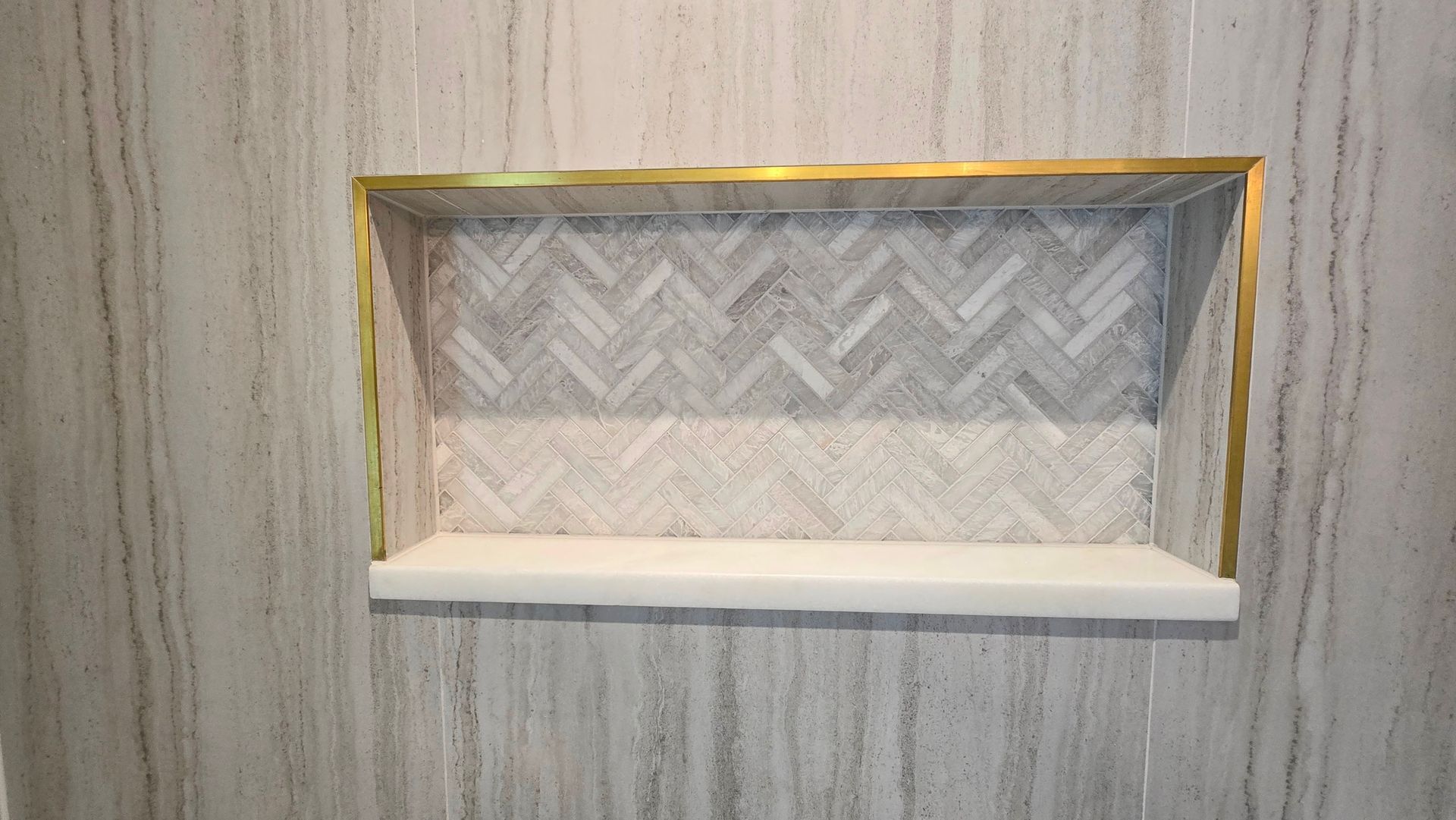 A shower wall niche in Cheshire, CT with light-veined marble tile, gold metal trim, herringbone-patterned interior tile, and a white ledge.