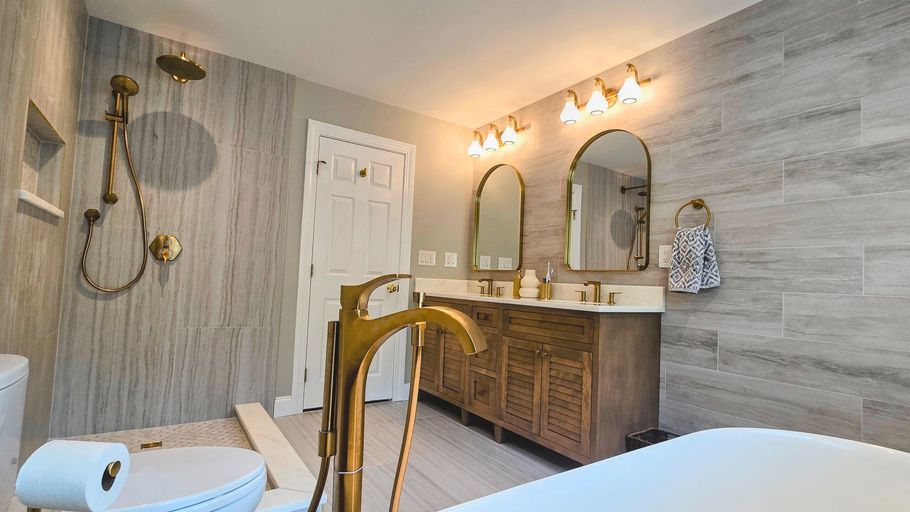 Bathroom in Prospect, CT with gold fixtures, wooden vanity, gray tile, white tub, and arched mirrors.