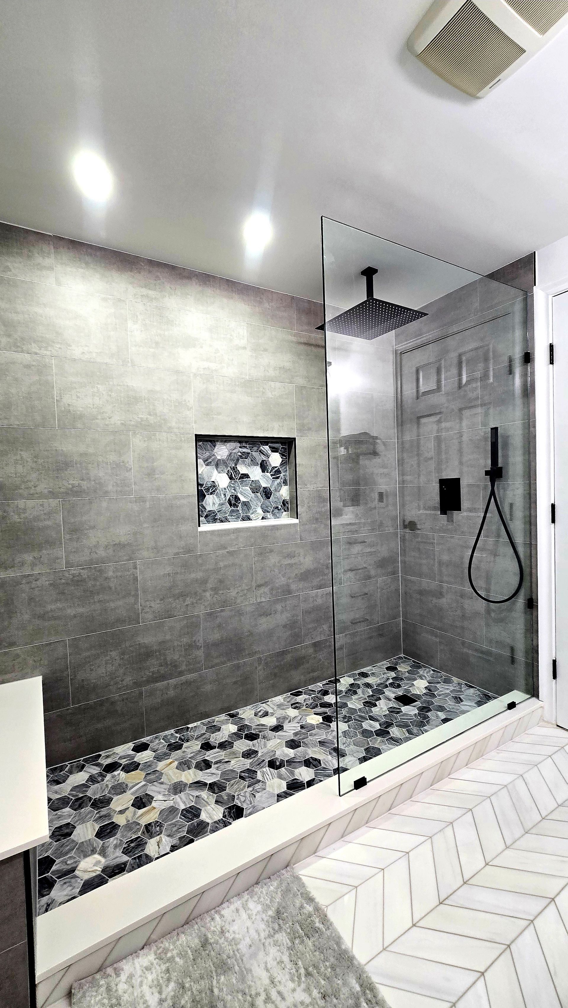 Modern walk-in shower in West Hartford, CT with gray tiled walls, pebble floor, rainfall showerhead, and a glass door in a bathroom.