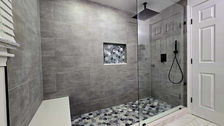 Gray-tiled shower in West Hartford, CT with pebble floor, glass door, and built-in shelf. Black showerhead and handheld wand. White door to the right.