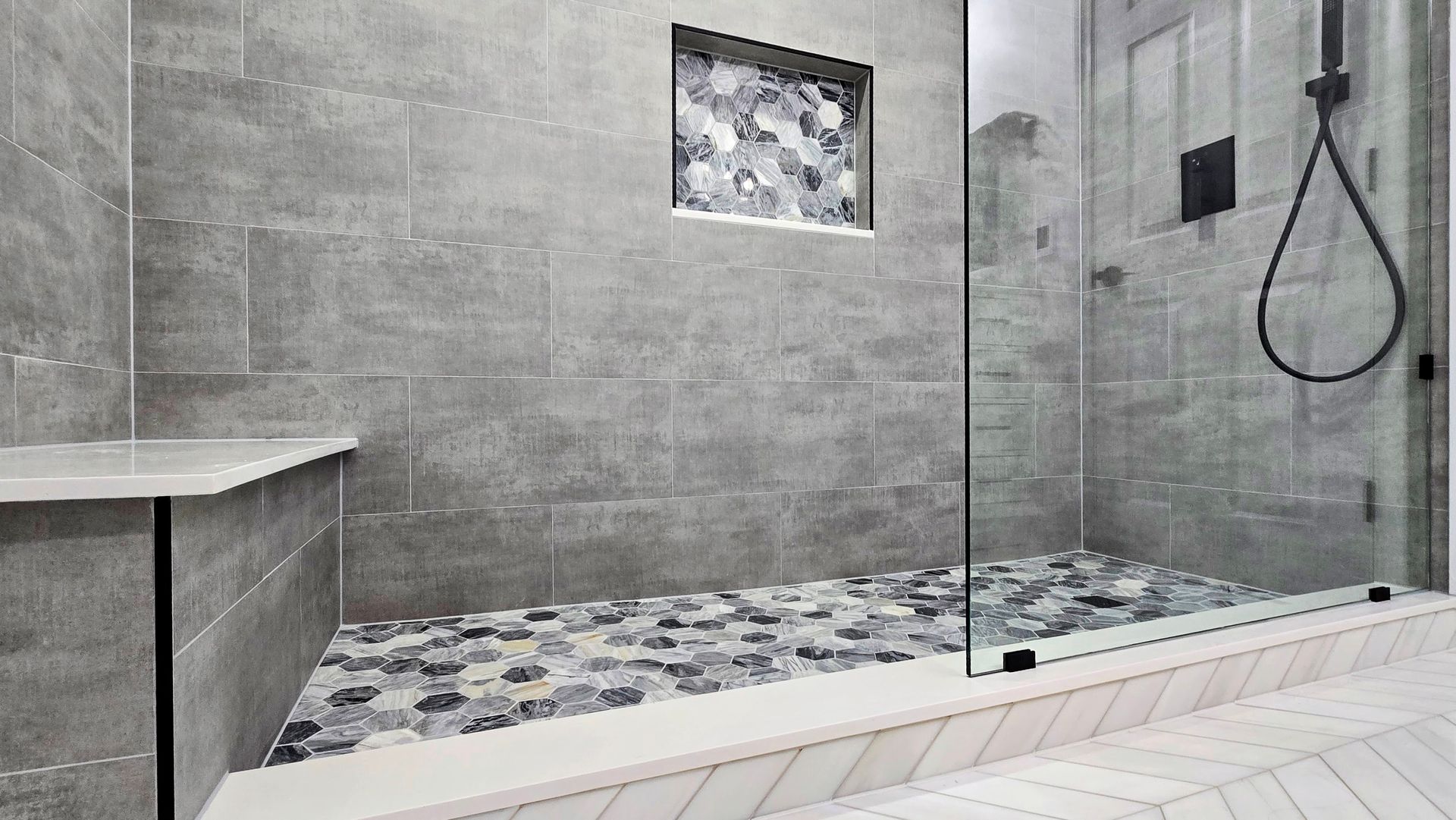 A modern walk-in shower in West Hartford, CT with grey tiled walls, a pebbled floor, a stone bench, and a glass partition.