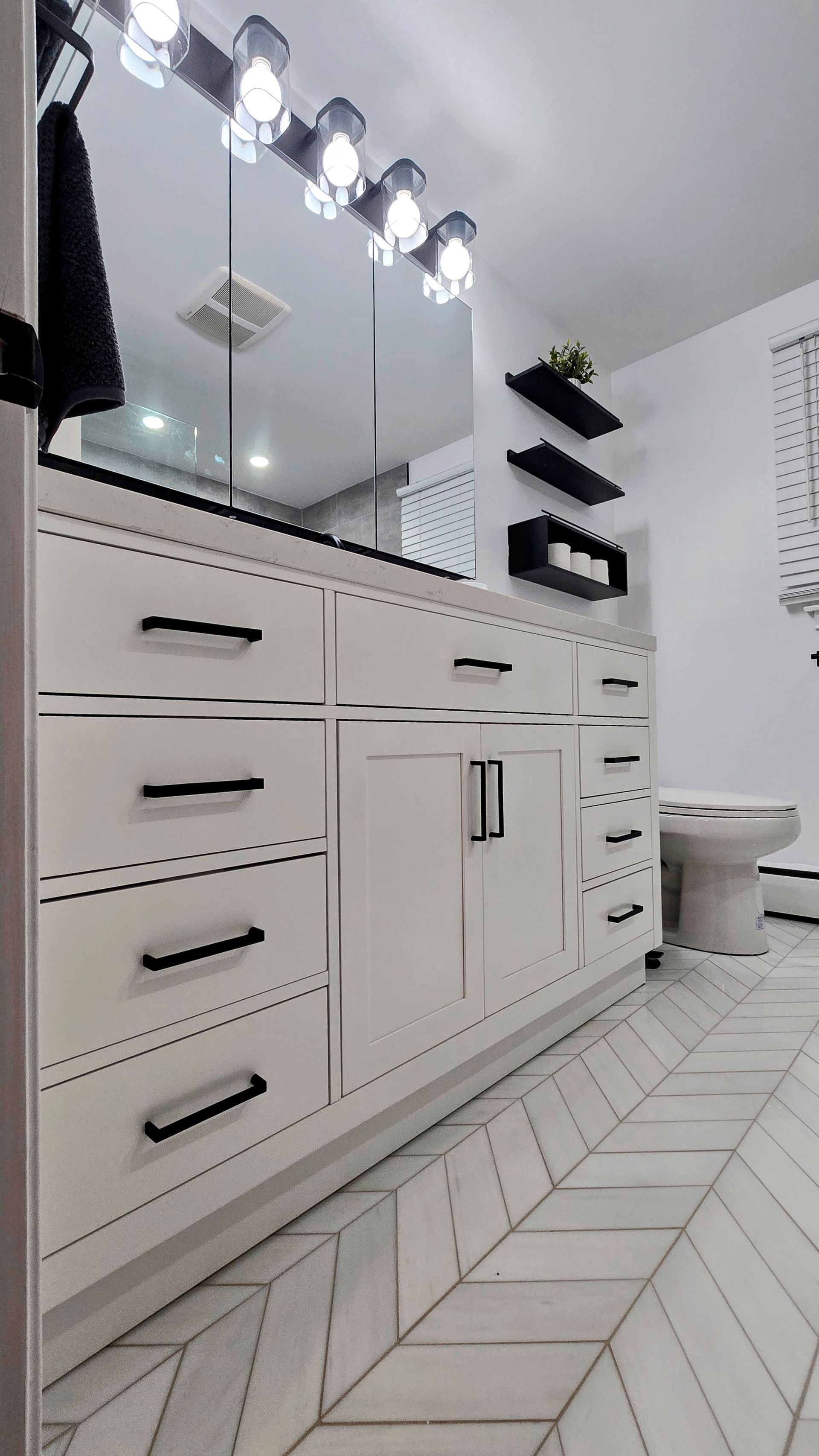 A bathroom vanity in West Hartford, CT with white cabinets, black hardware, a lit mirror, and light wood herringbone tile floors.