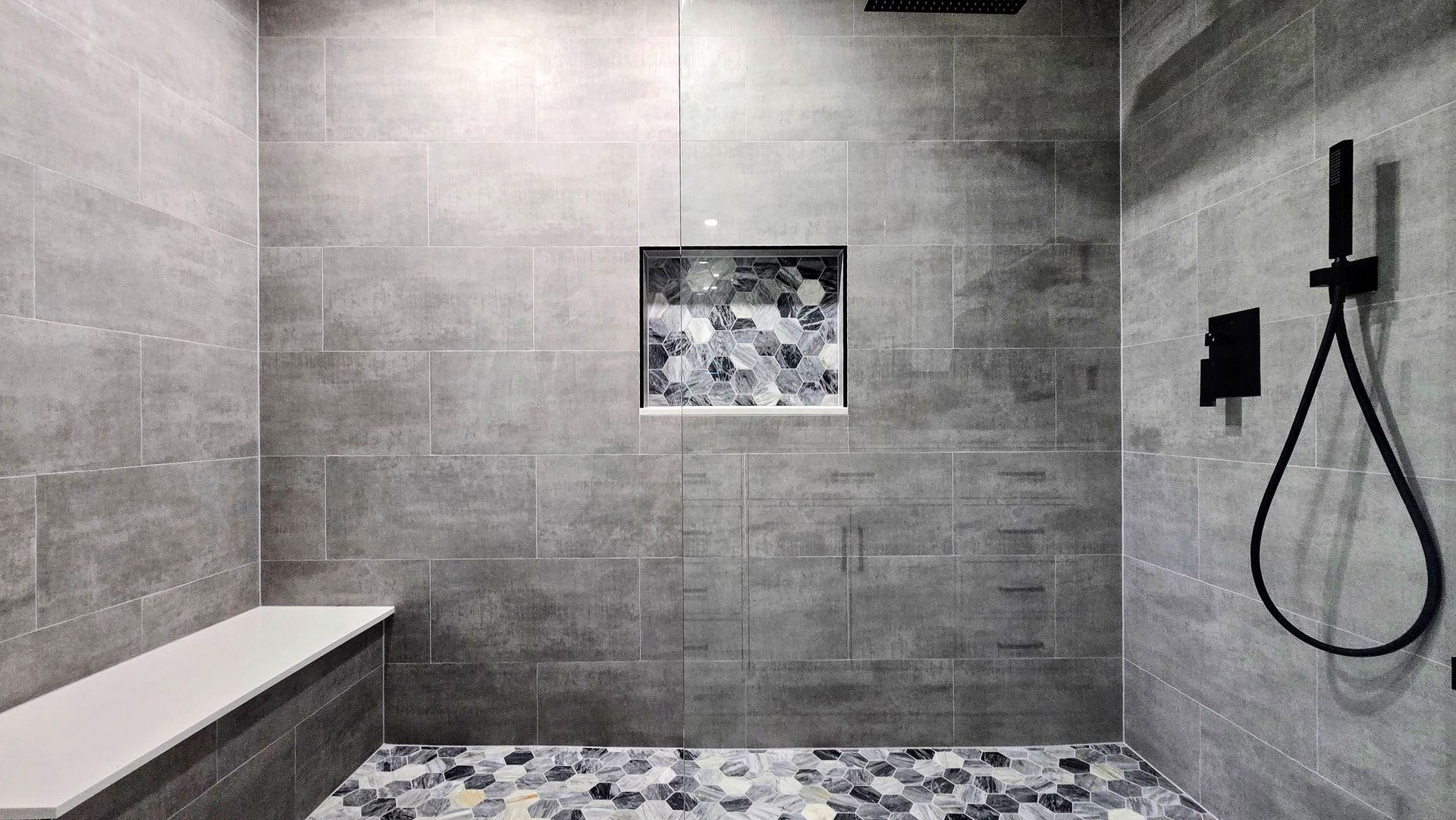 A walk-in shower remodel in West Hartford, CT with gray tiled walls, a pebble floor, a built-in bench, and a black handheld shower fixture.