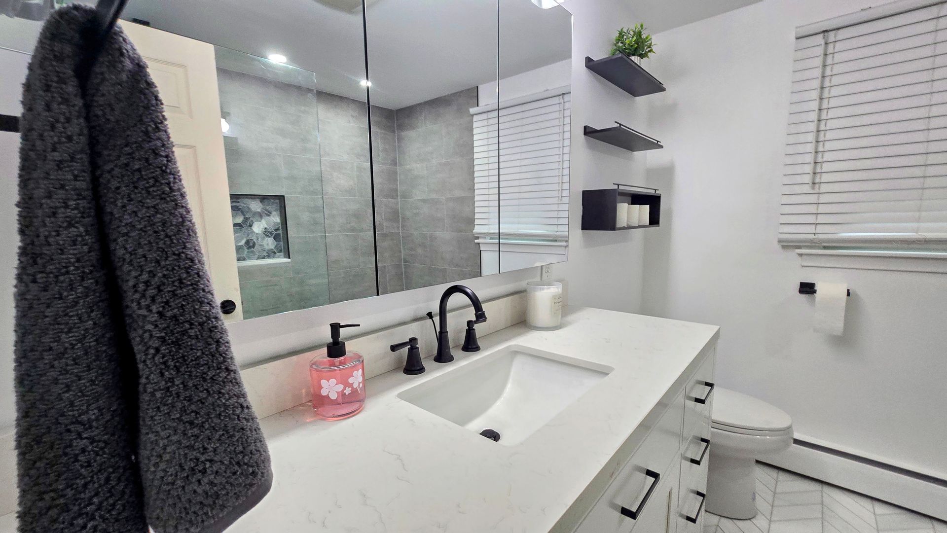 A modern, white-toned bathroom remodel in West Hartford, CT featuring a stone countertop, a square sink, a large mirror, and open shelving.