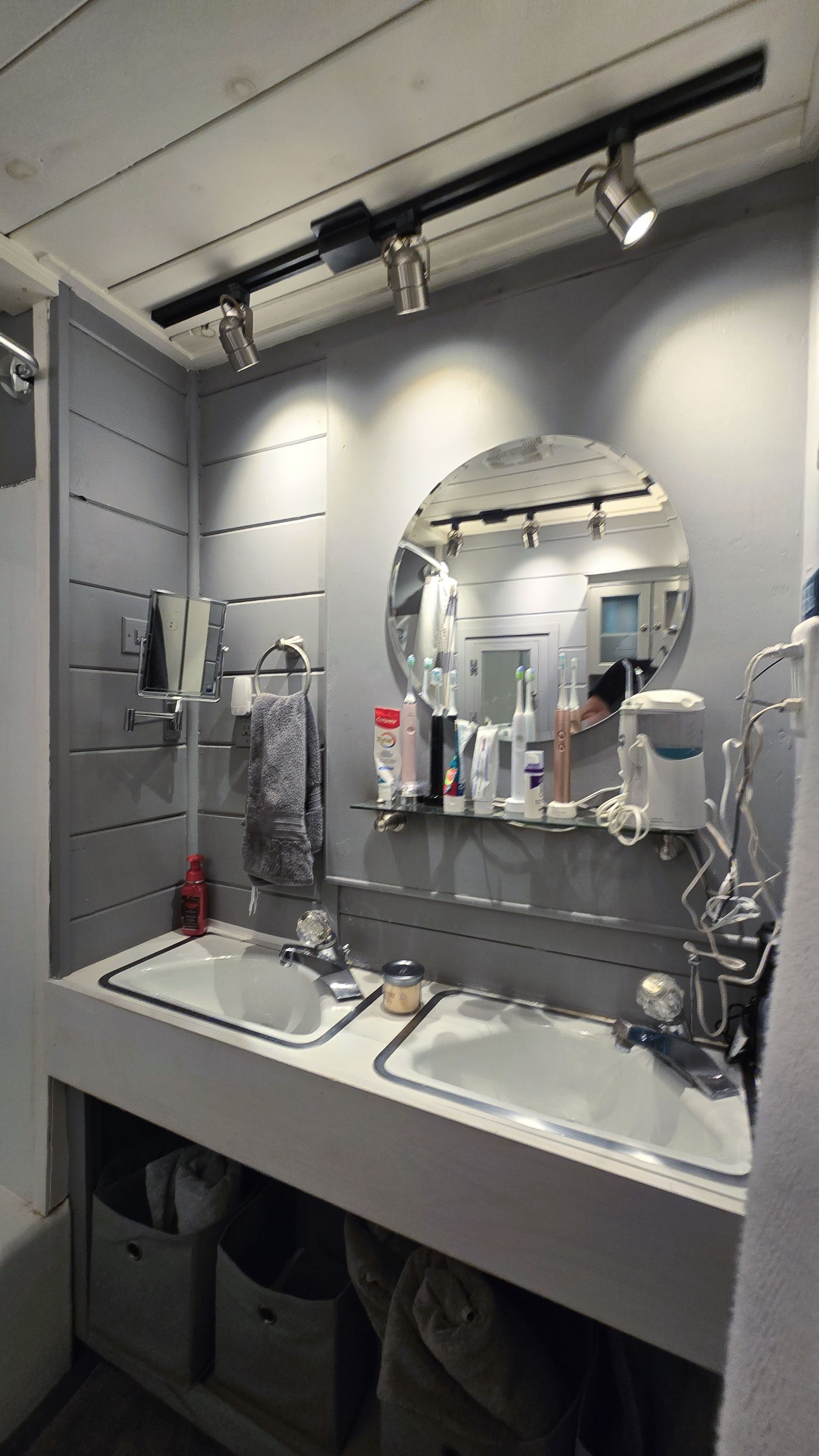 Bathroom with double sinks, round mirror, shelf with toiletries, track lighting. Light gray walls and cabinets.