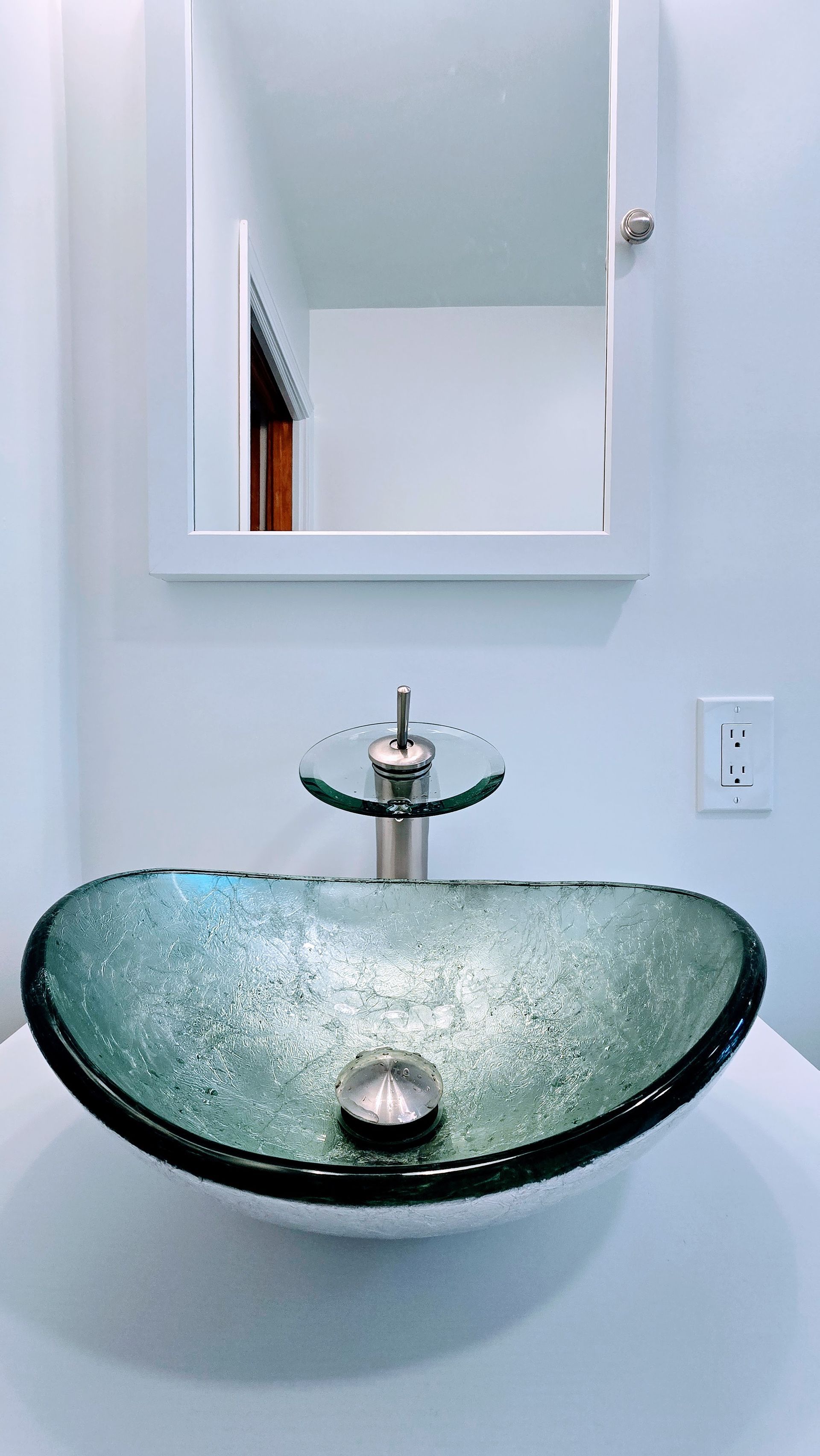 Oval, teal-colored glass vessel sink with brushed nickel faucet, mounted on a white countertop in a modern bathroom. A white medicine cabinet hangs above.