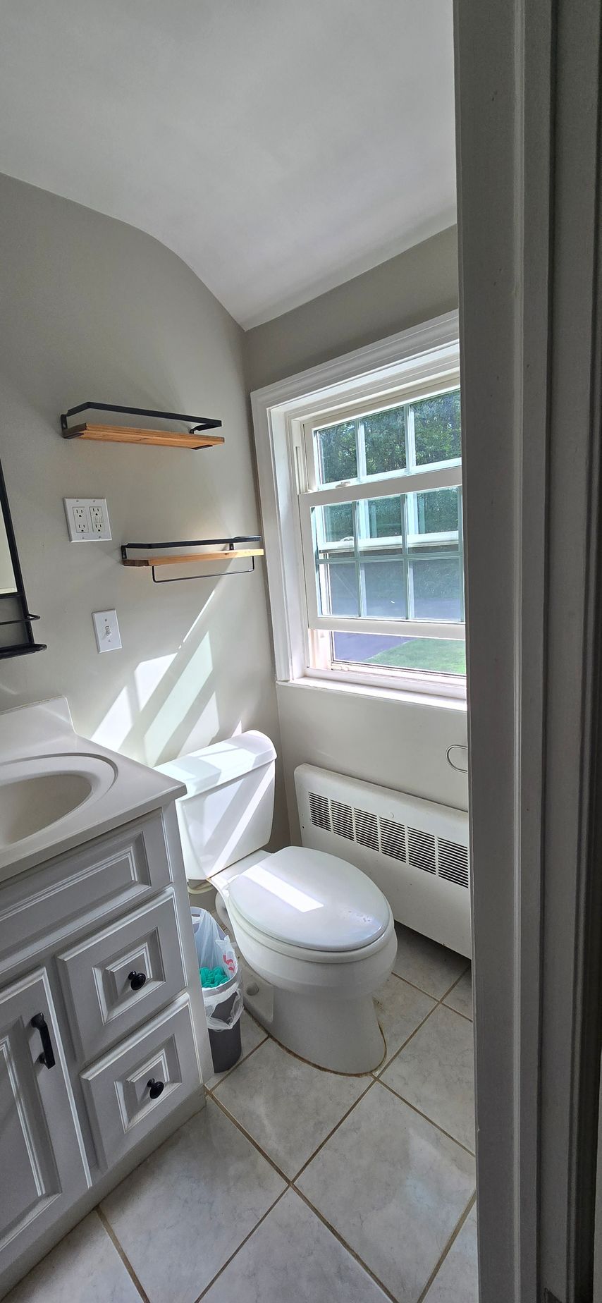A small, well-lit bathroom with a white toilet, vanity, and a window with a view.