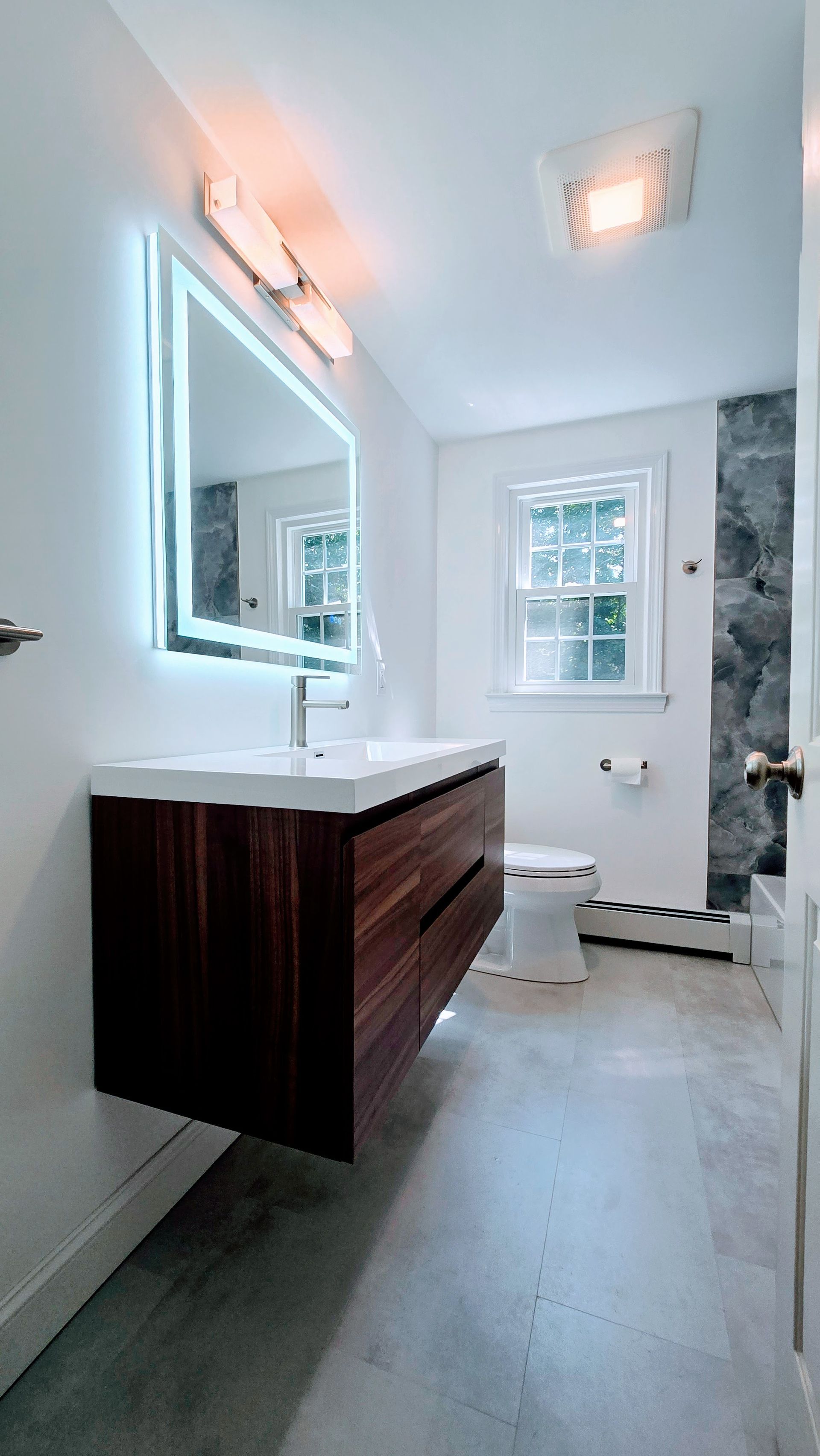 Modern bathroom with floating wood vanity, illuminated mirror, and small window.