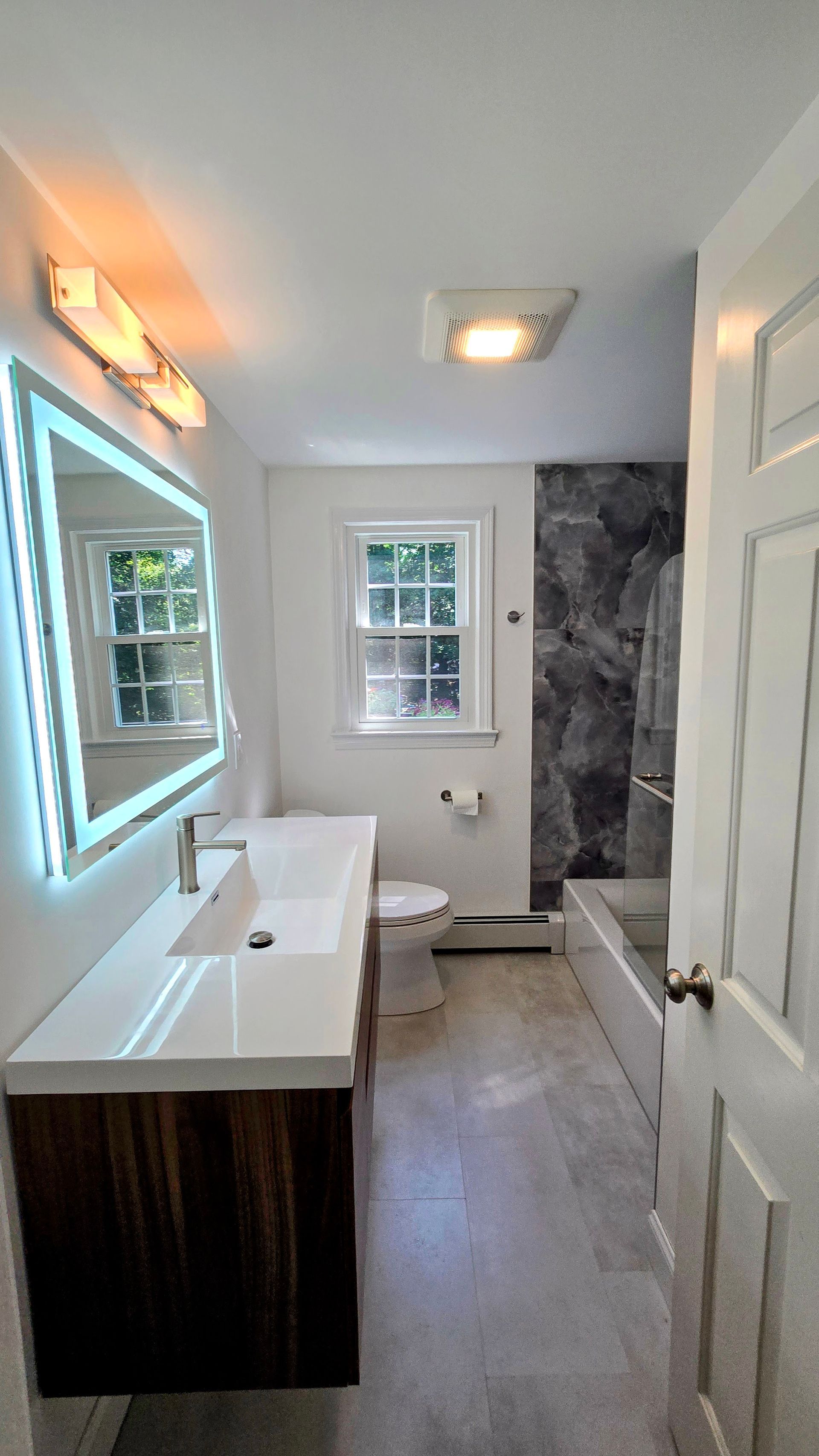 Modern bathroom with white walls, wood vanity, and a gray marble accent wall. A lit mirror hangs over the sink, and a window is centered on the back wall.