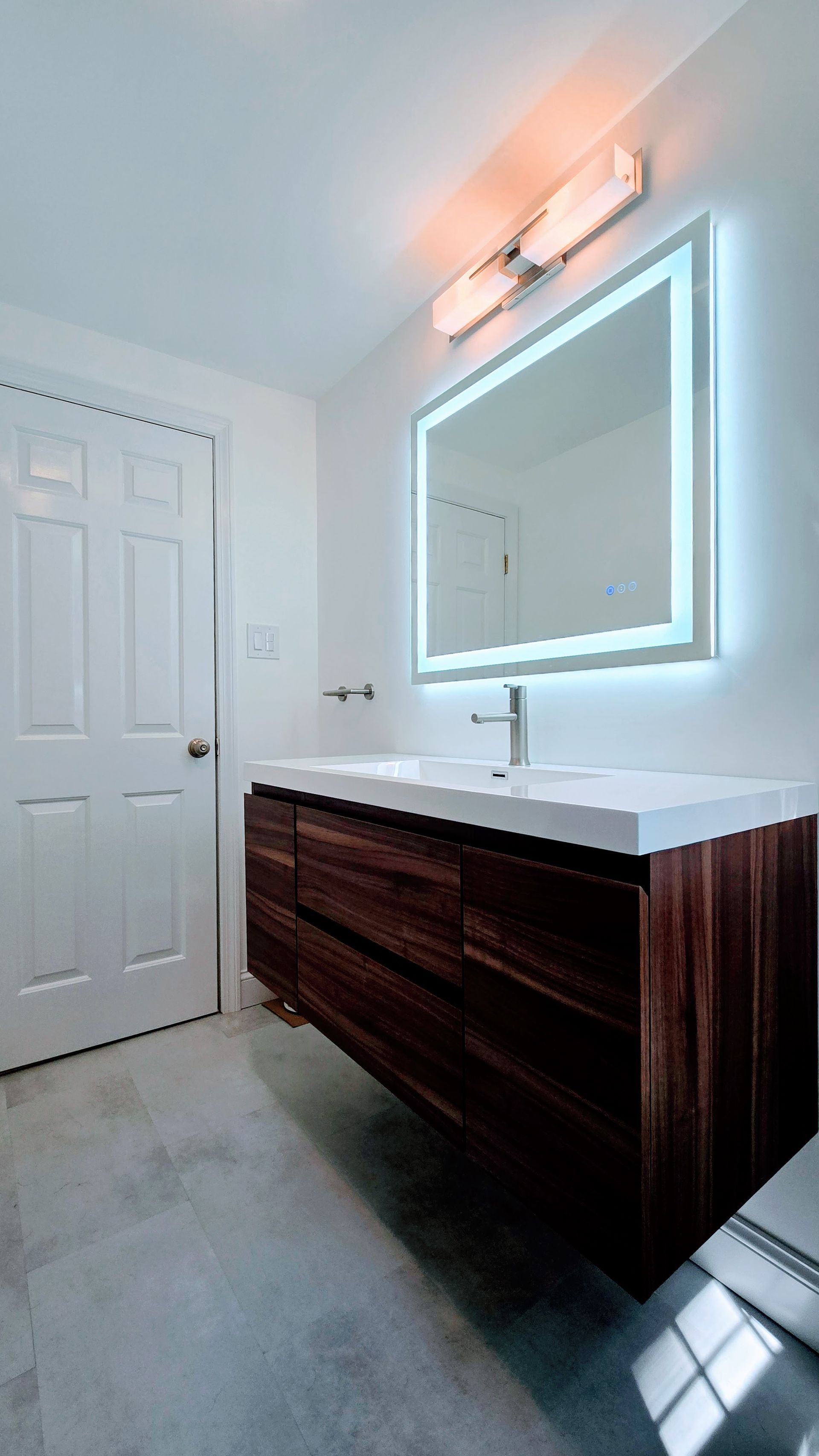 Modern bathroom with floating wood vanity, backlit mirror, and gray tile floor.
