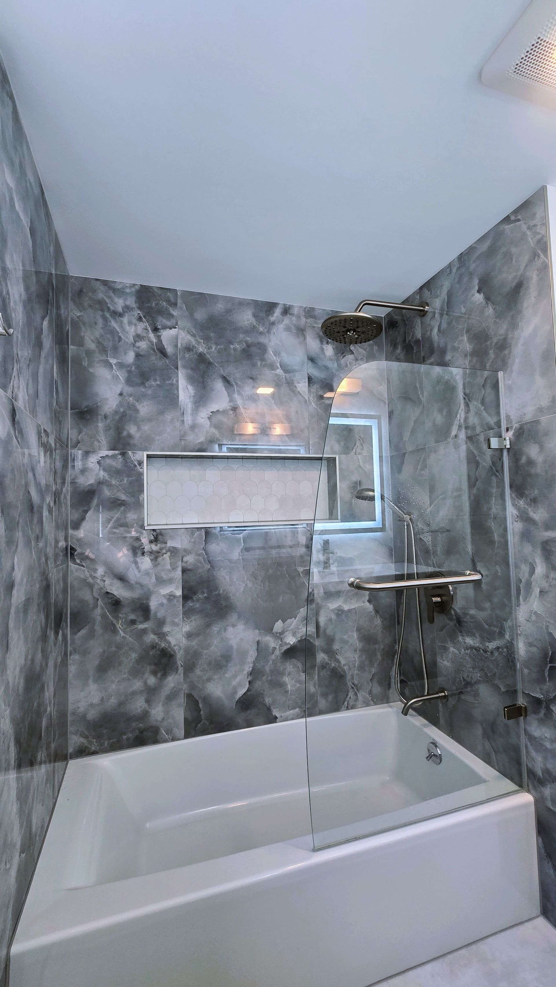 A modern bathroom with a white bathtub, gray marble-look tiled shower walls, a glass shower door, and built-in shelf.