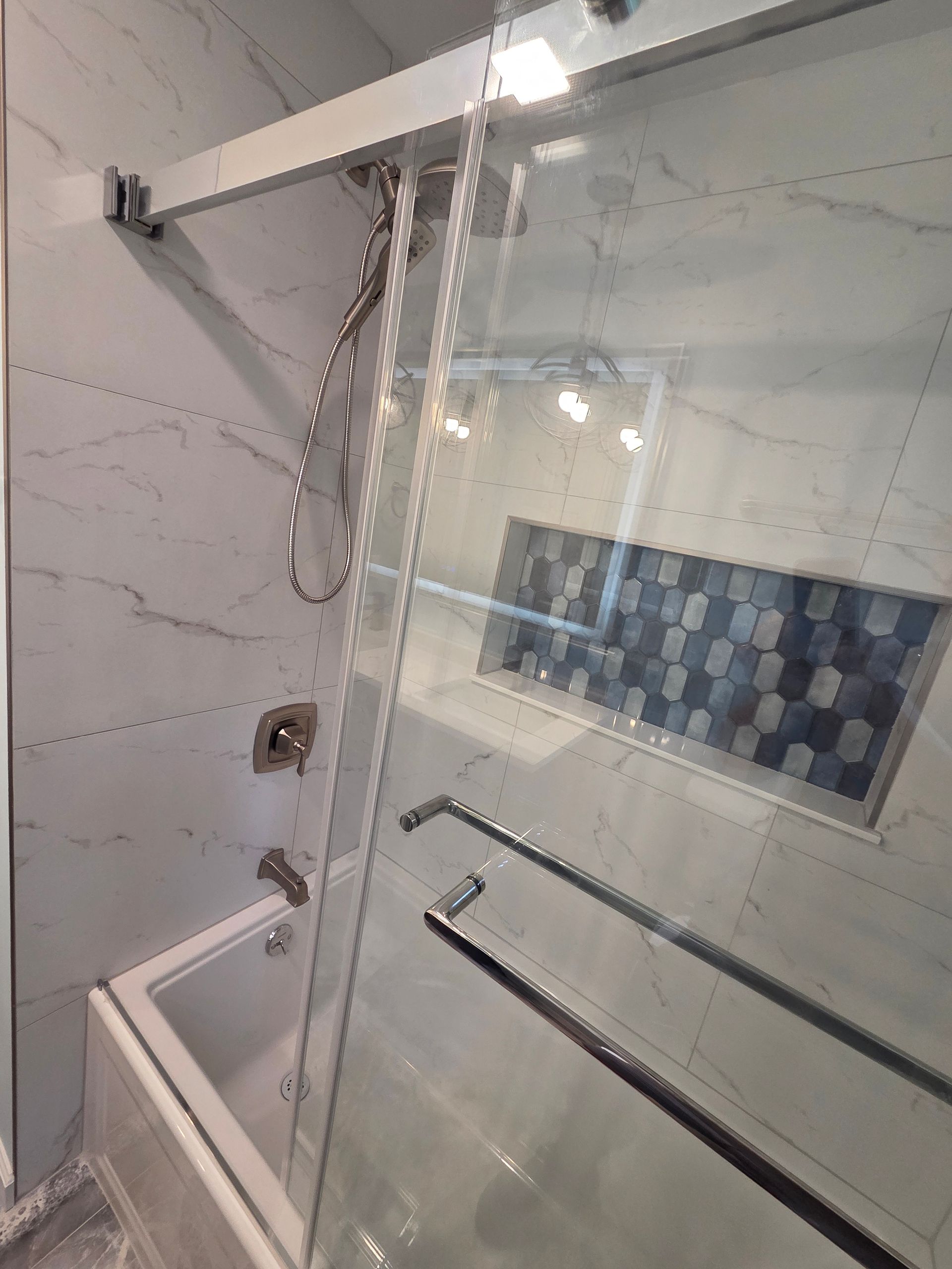 Shower with glass door, chrome fixtures, white marble tile. Built-in shelf with blue mosaic tile detail.