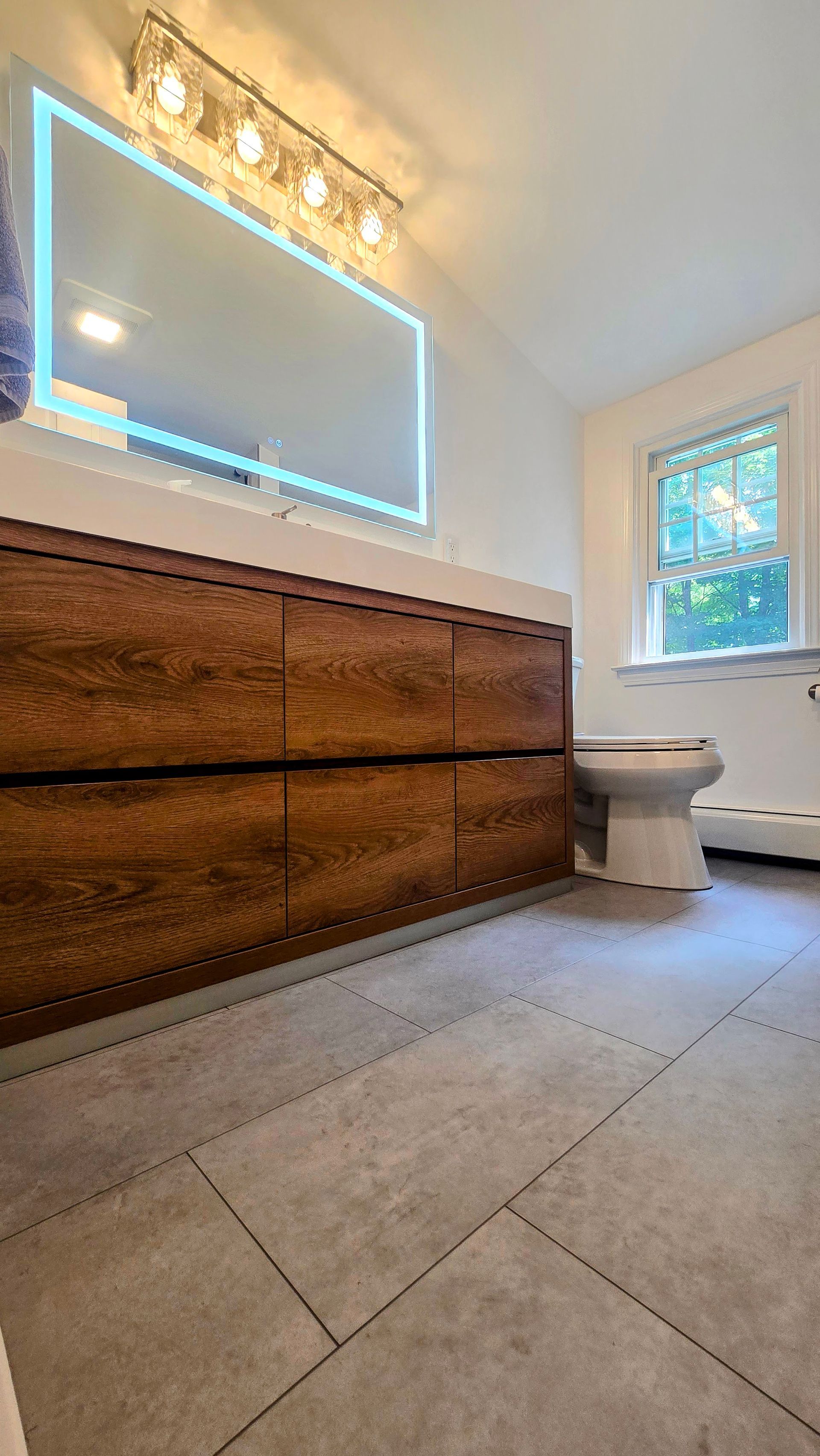 A modern bathroom with a wood vanity, large mirror, and gray tile floor. A toilet and window are also visible.