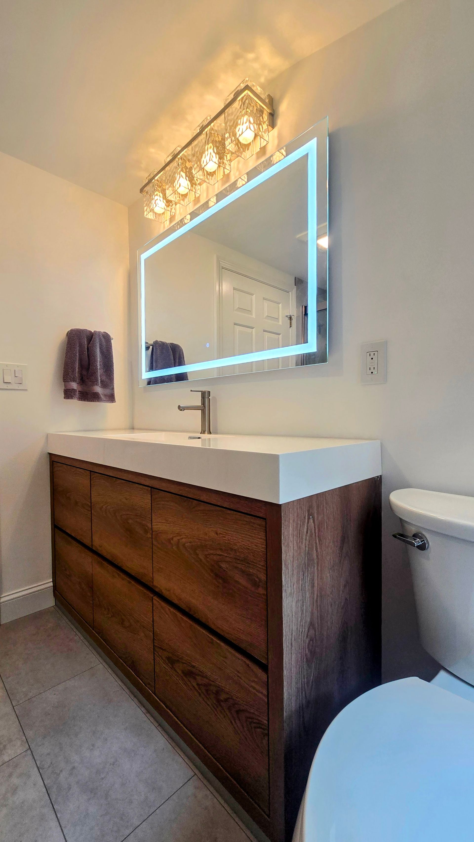 Modern bathroom with a brown wooden vanity, rectangular lit mirror, and a toilet.