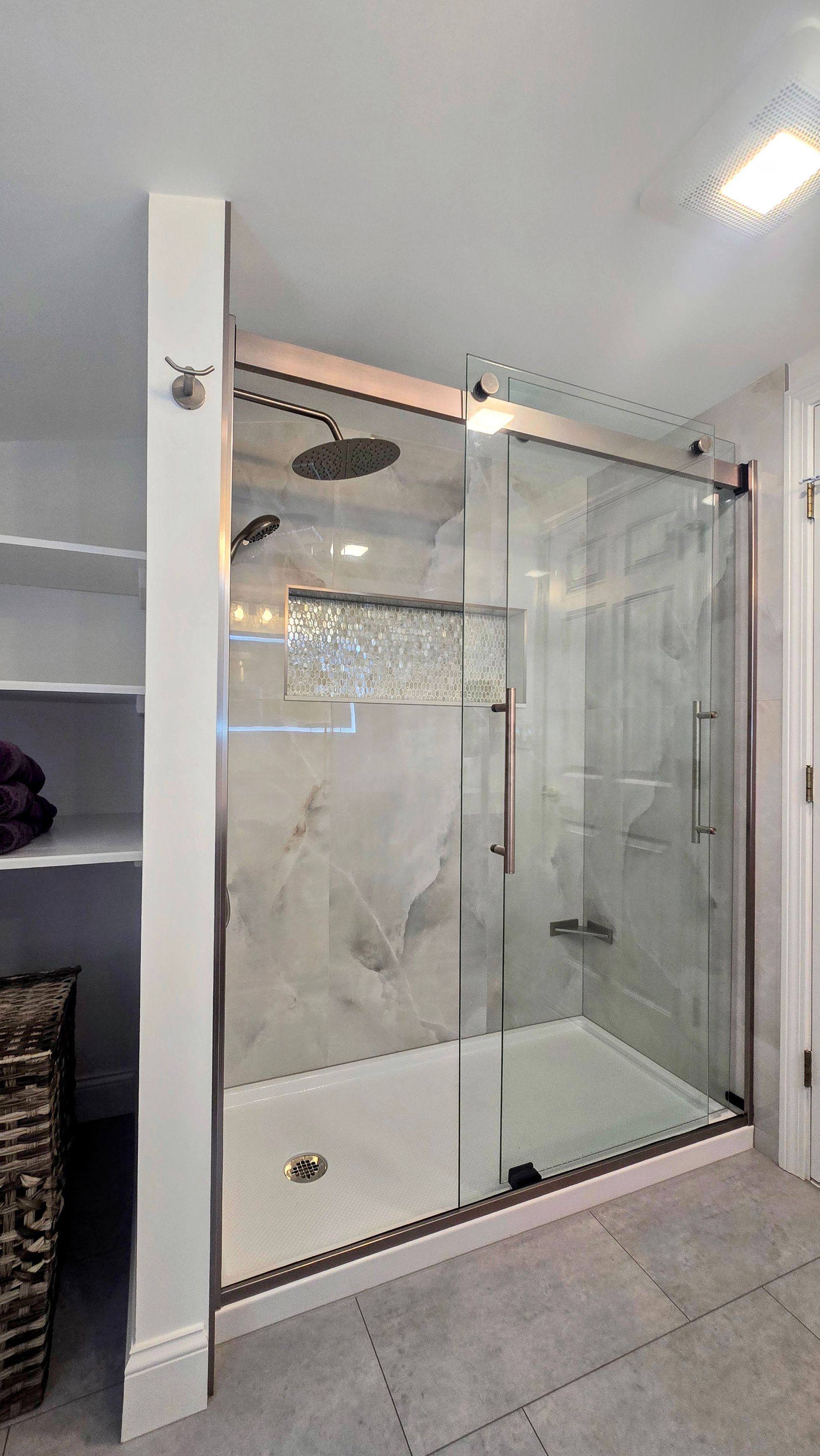 Modern shower with glass doors, white base, gray tile floor. Next to a shelving unit with basket.