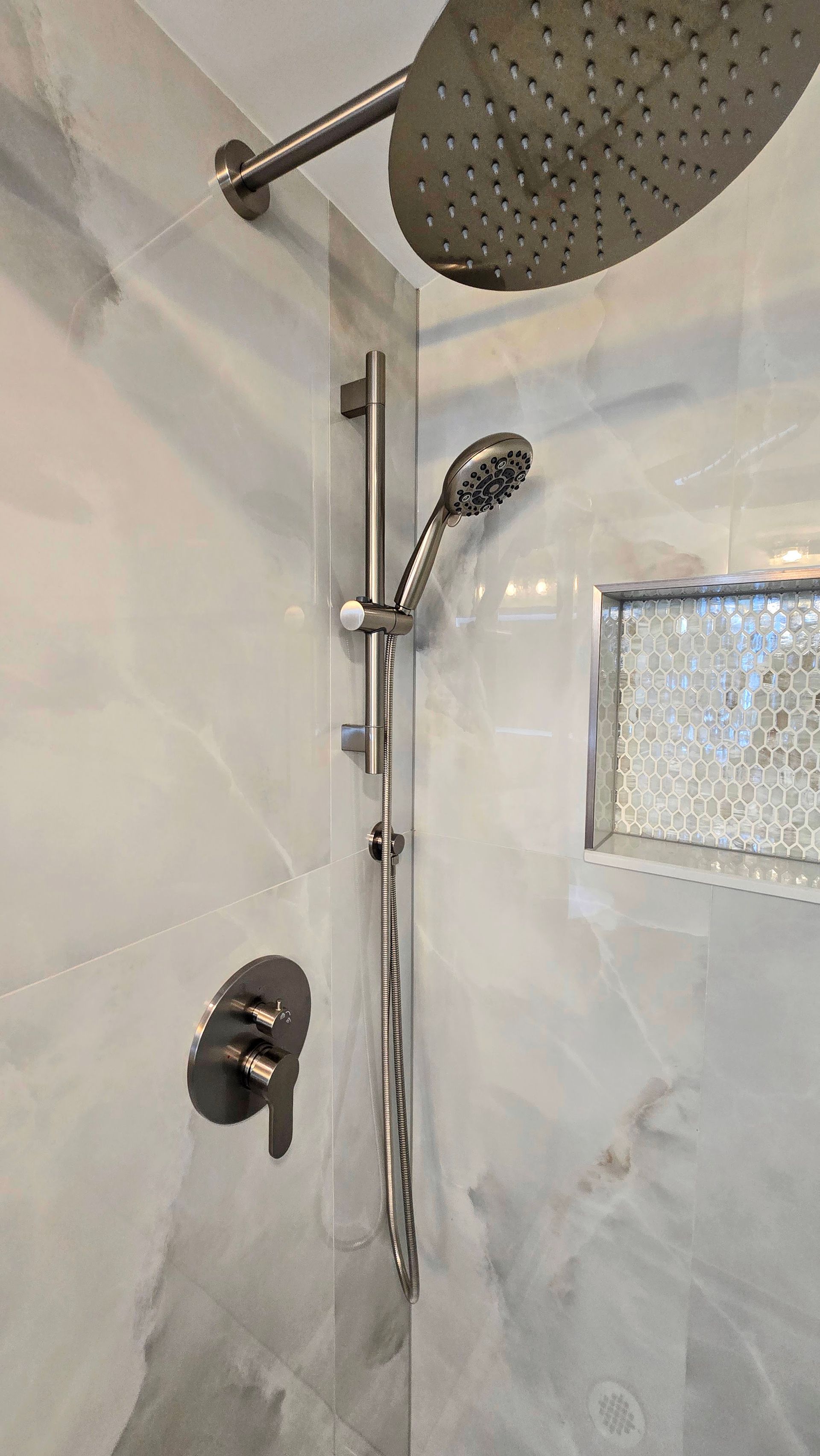 Shower with rain head, hand shower on adjustable bar, and wall-mounted faucet on marble-like tile.