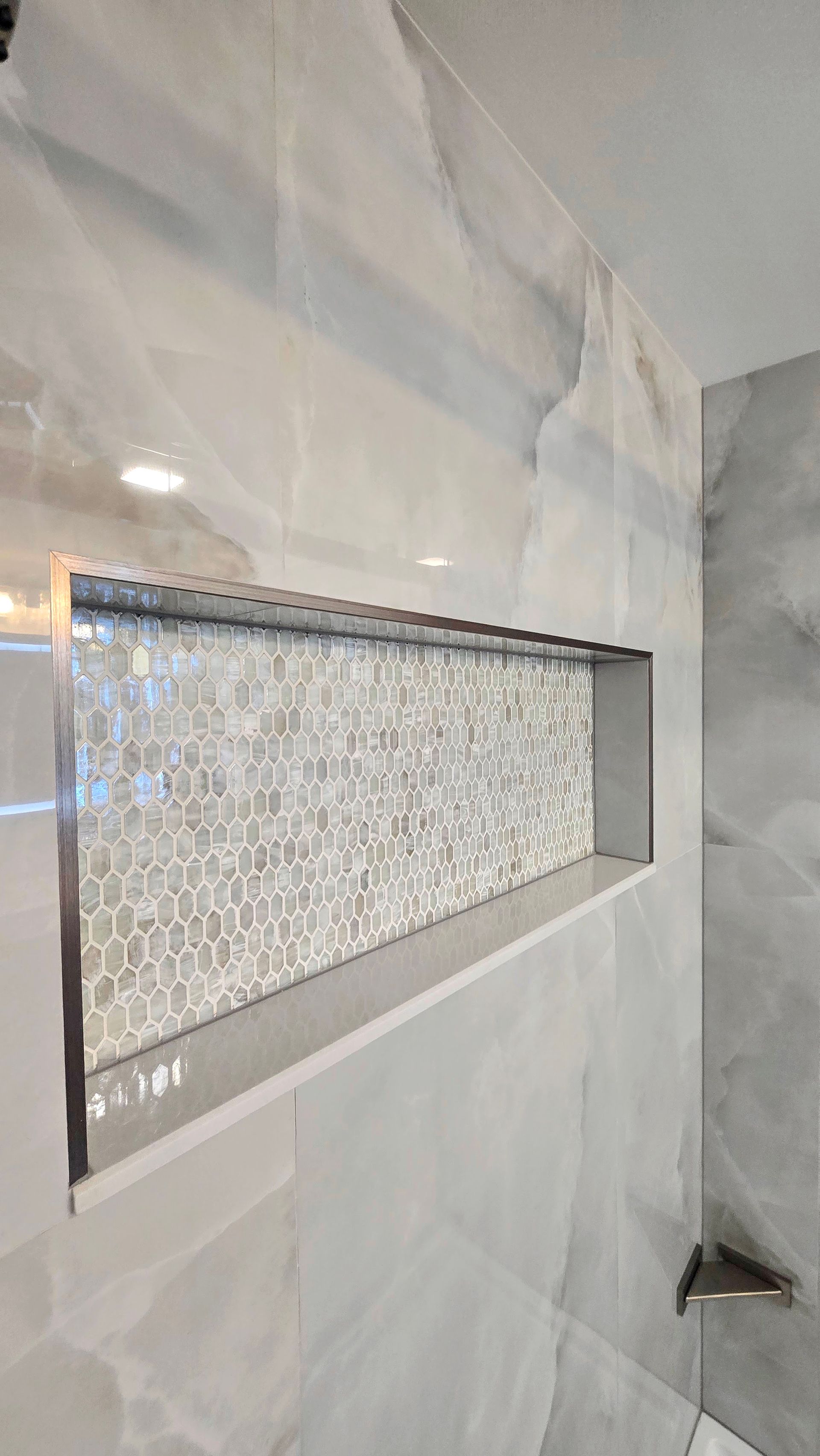 Shower niche with small, mosaic tile back and surrounding large, marble-look tiles. The niche has a metal frame.
