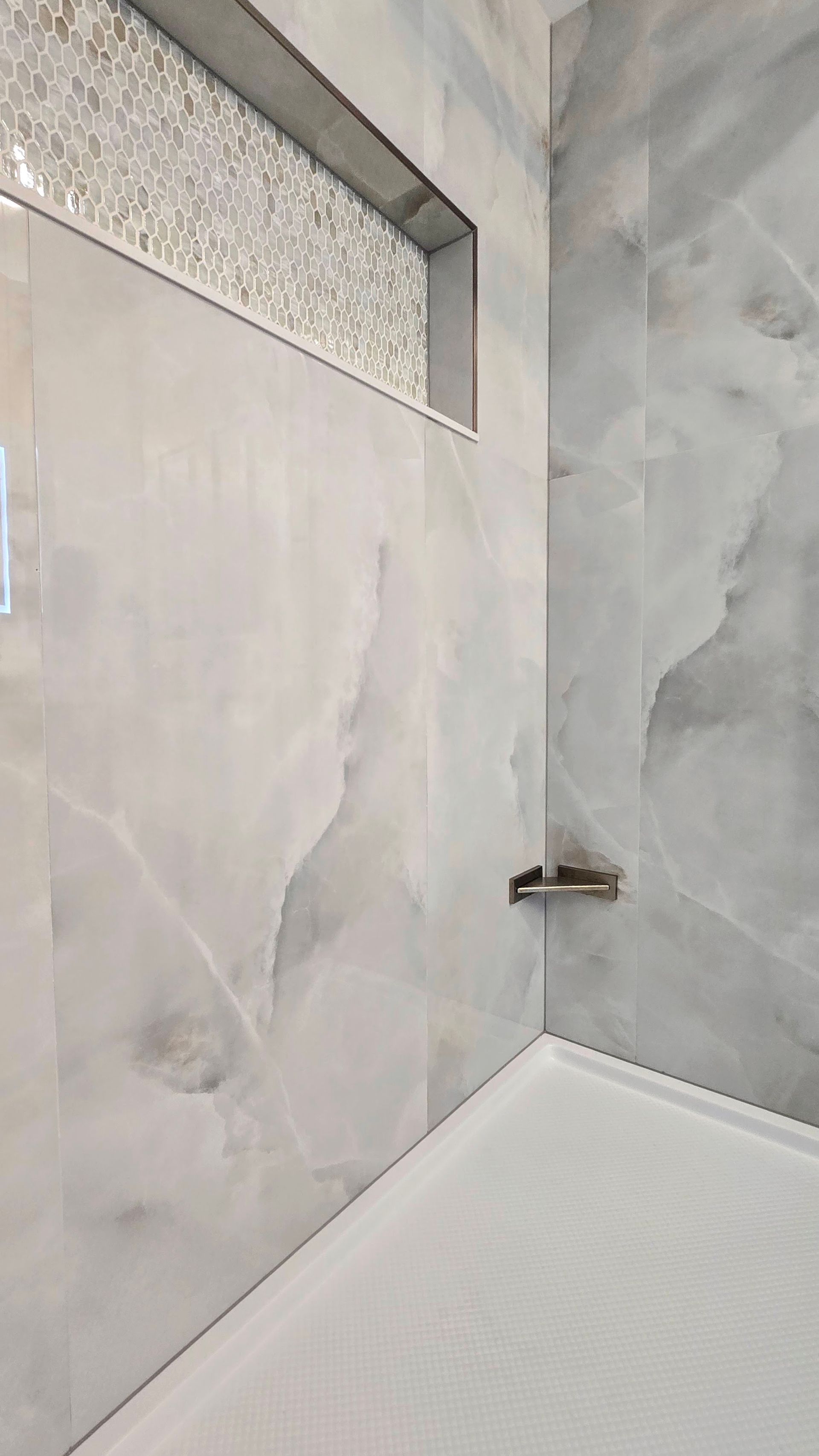 Close-up of a modern shower with large, light gray marble-look tiles, a small shelf, and a recessed mosaic tile accent.
