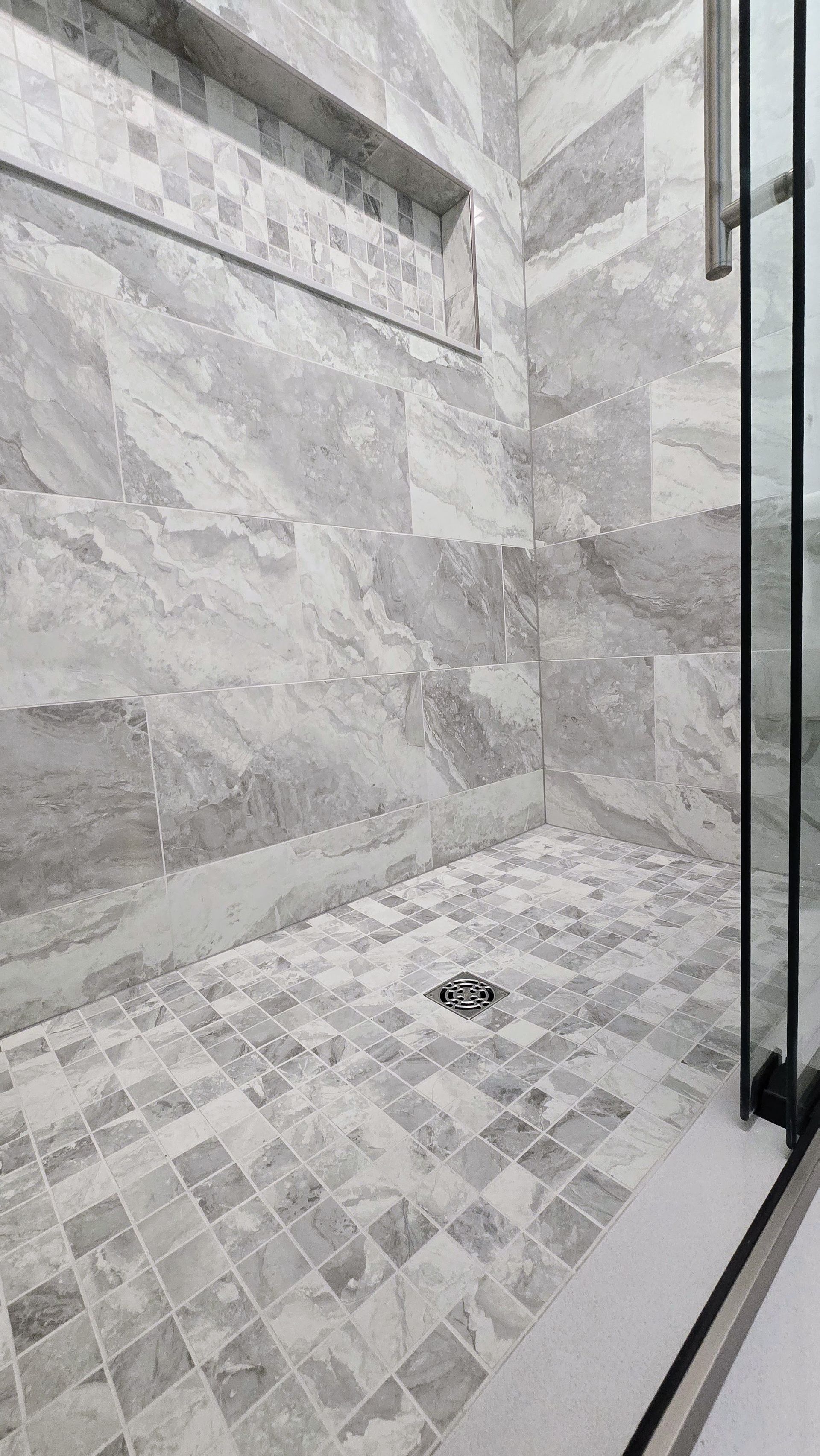 Gray tiled shower with glass door and built-in shelf. Mosaic tile floor.