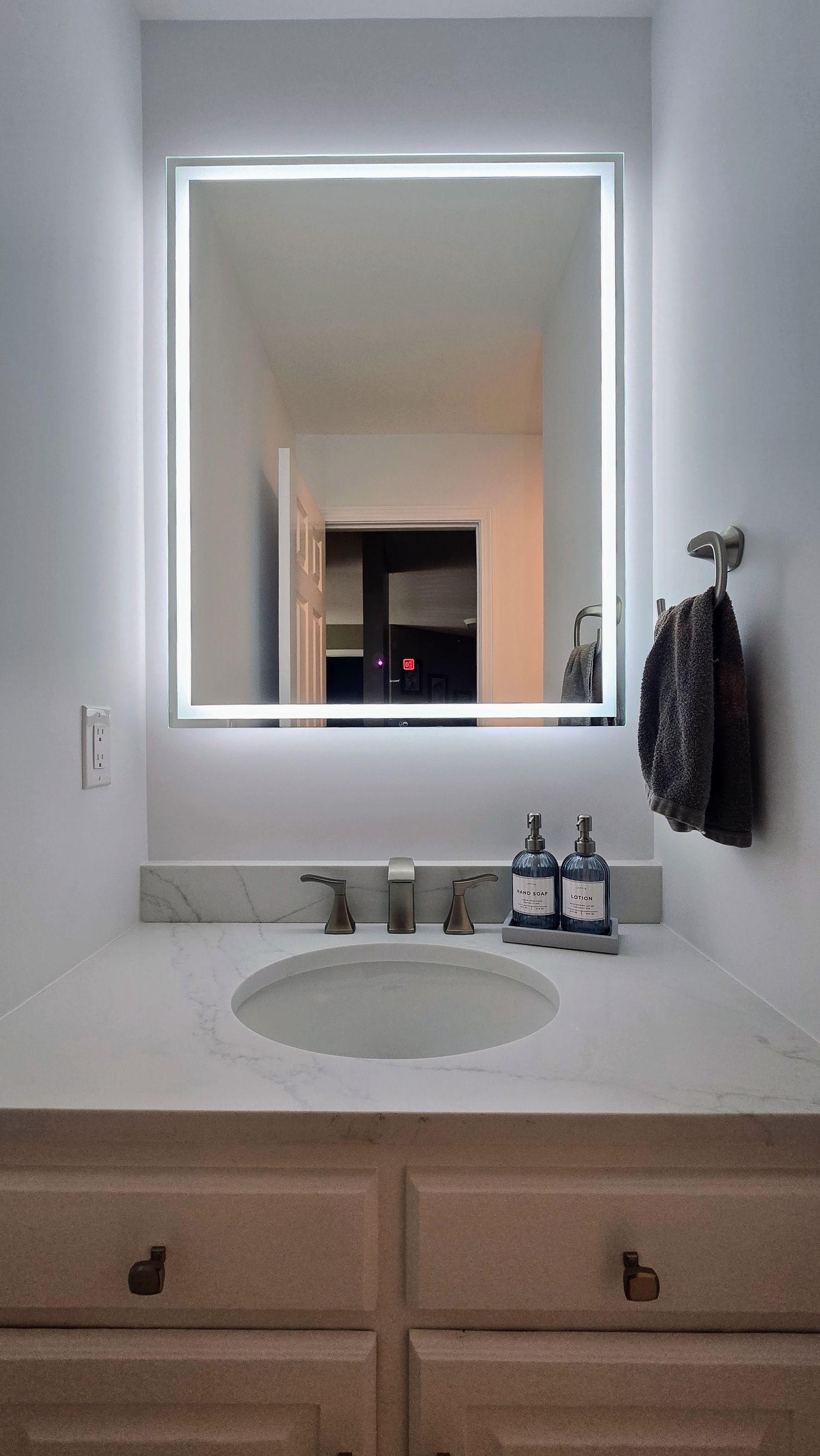 Bathroom with lit mirror, marble countertop, white cabinets, and a sink.