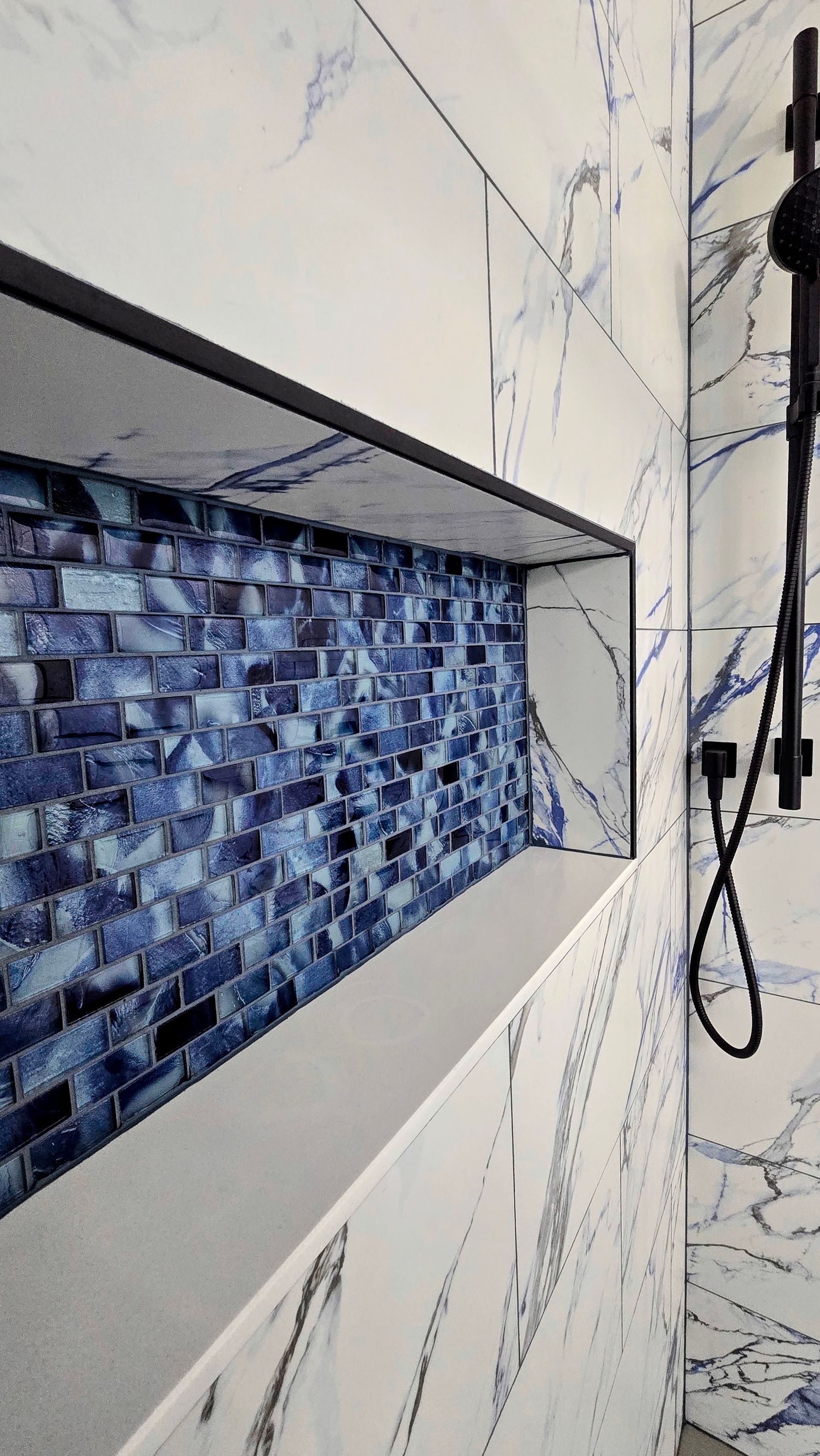 Shower niche with blue mosaic tile and marble-patterned white tile. Black shower fixtures on the right.