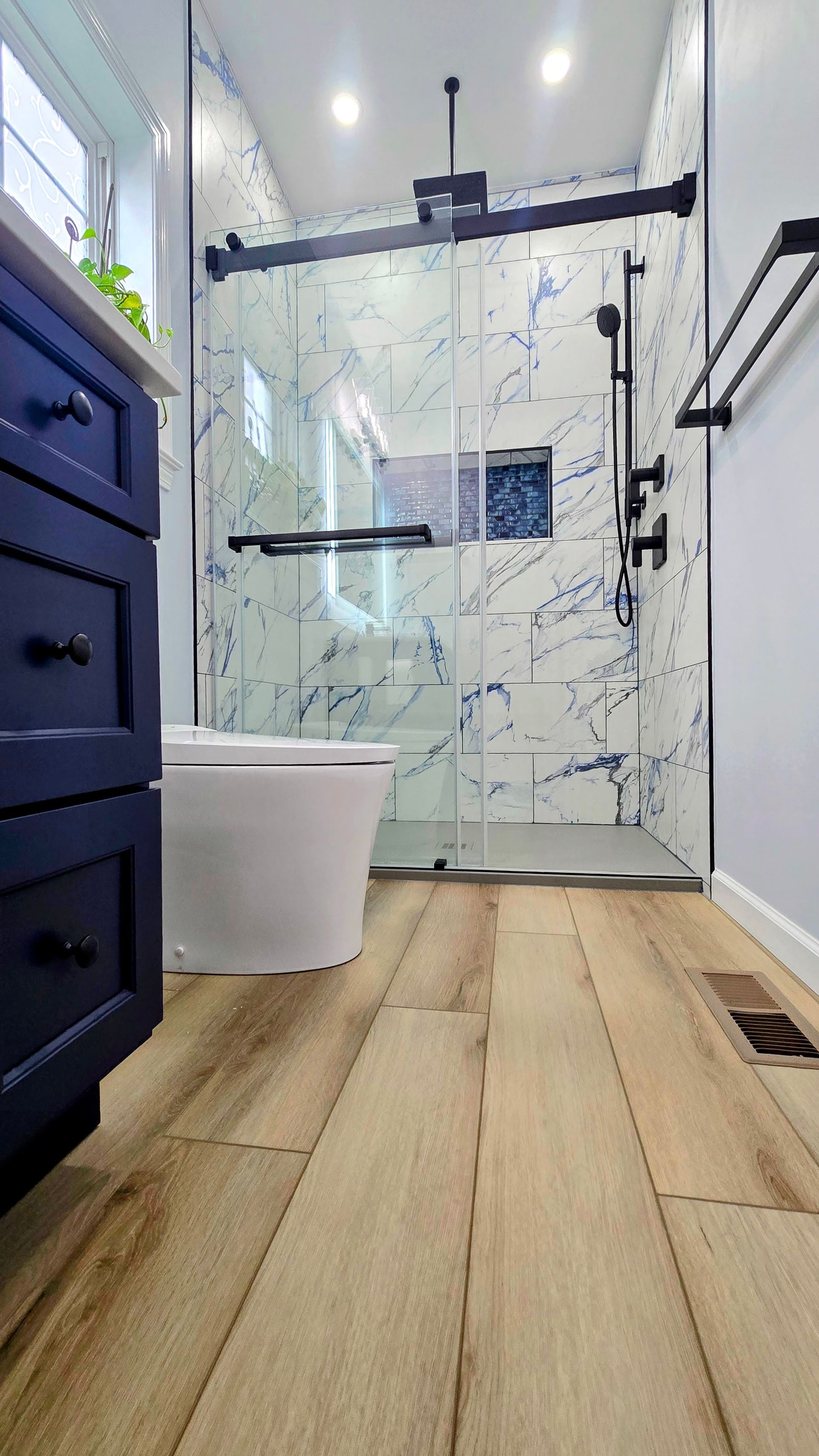 Bathroom with blue vanity, white toilet, glass shower, and wood-look tile.