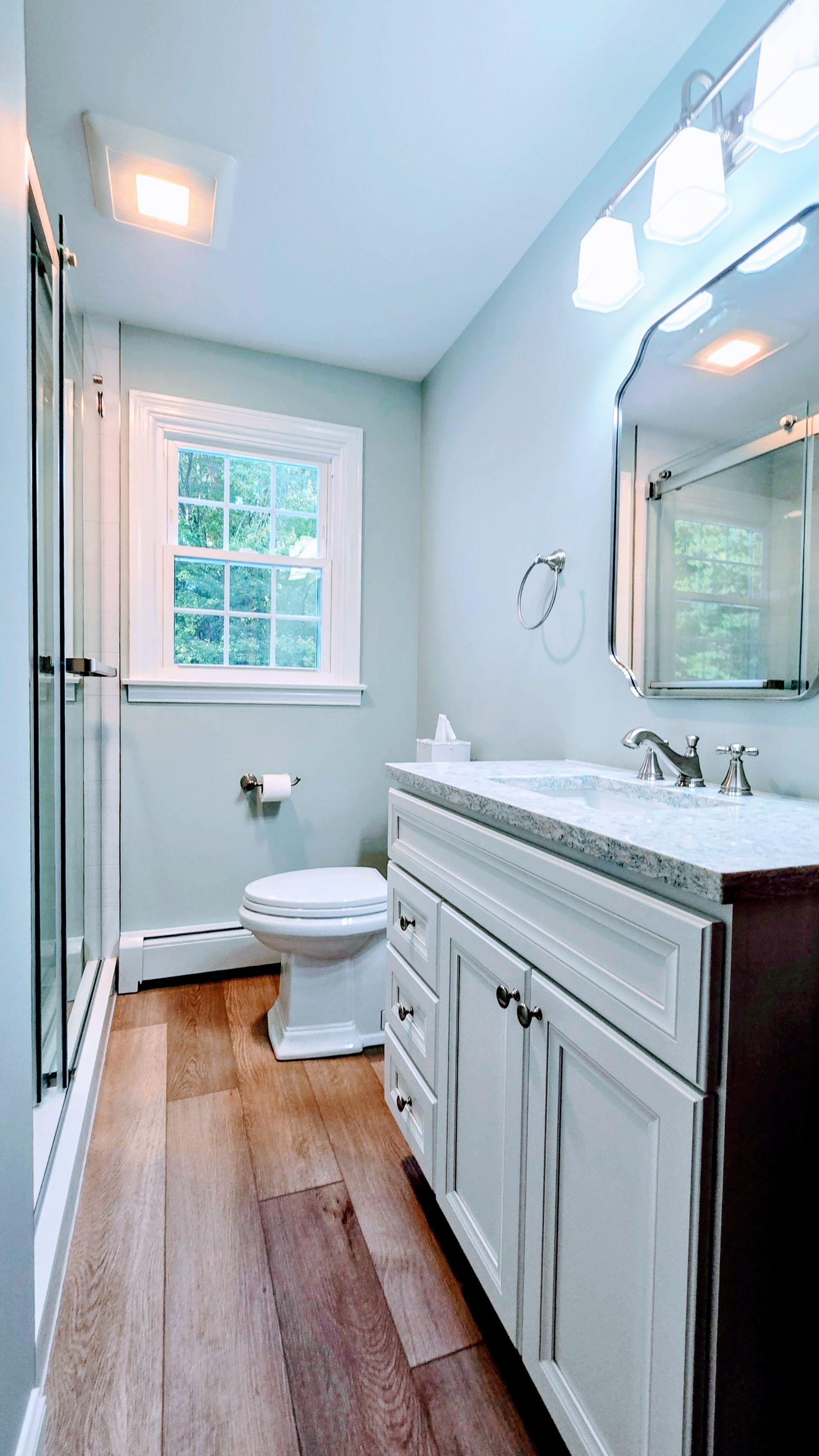Bathroom remodel in Bristol, CT with white vanity, toilet, and window. Light blue walls, hardwood floor.