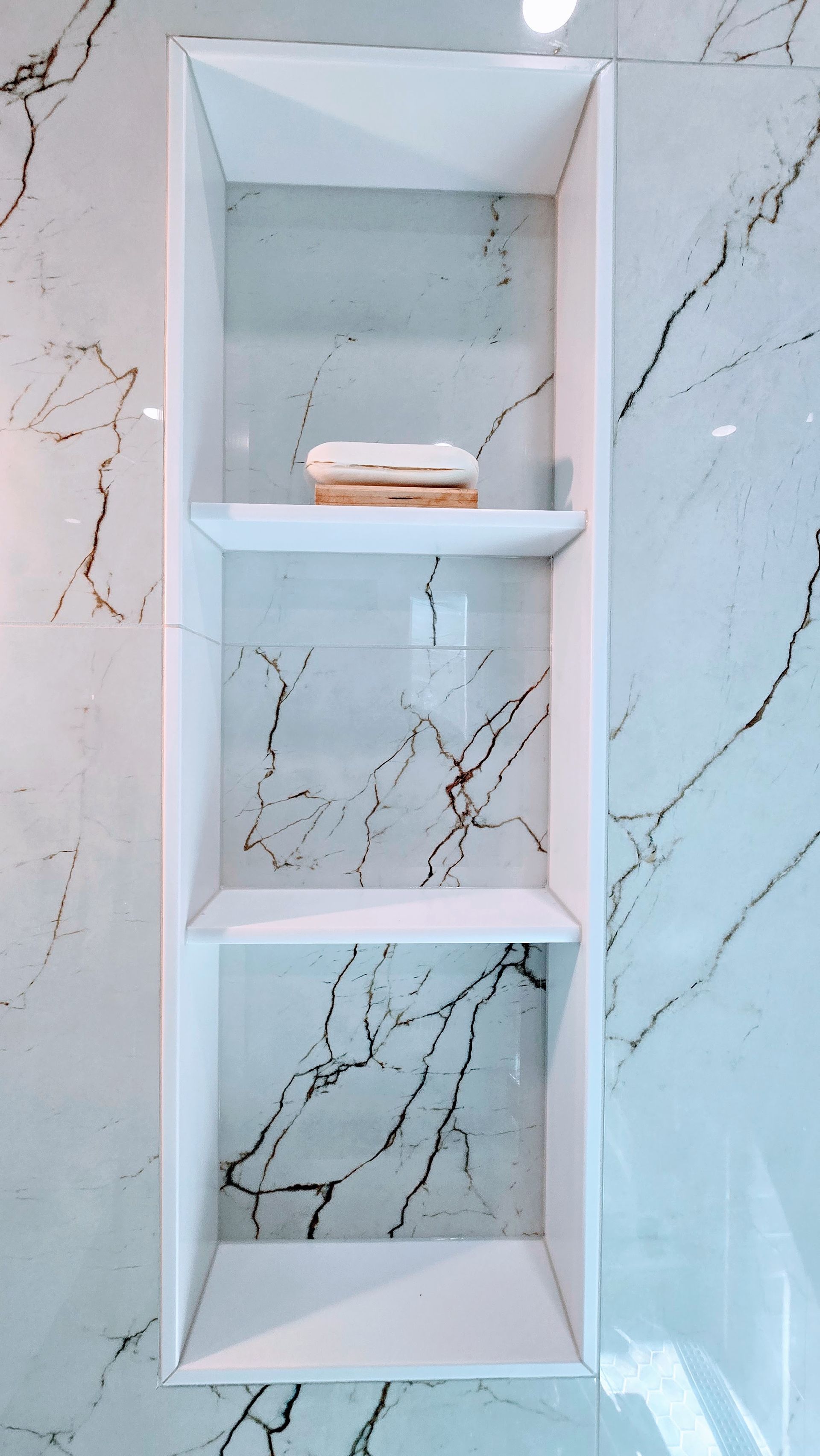 White built-in shelf with three sections in marble-tiled wall. A bar of soap rests on middle shelf.