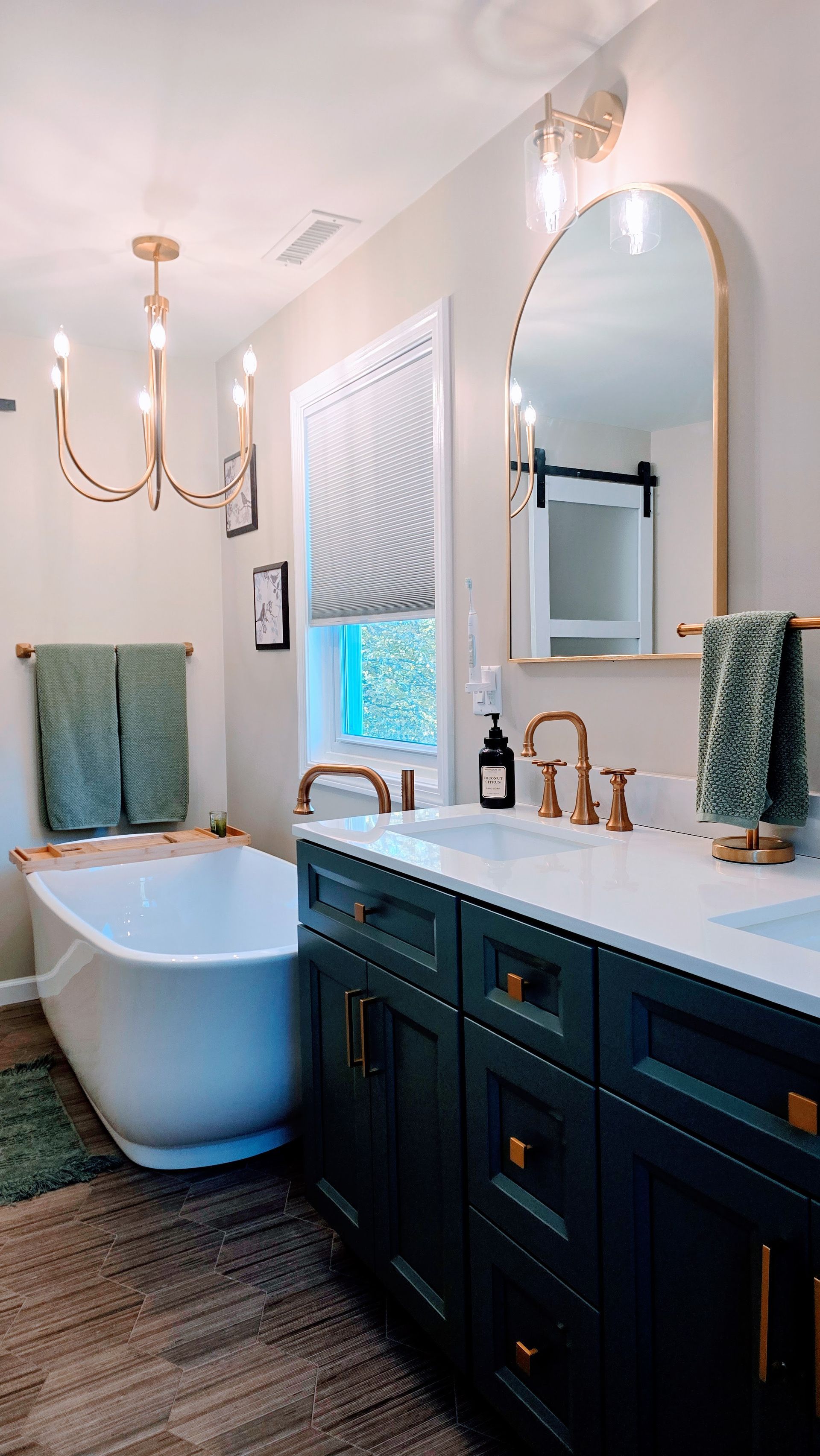 Elegant bathroom in Wolcott, CT with a dark green vanity, white tub, gold fixtures, and arched mirror.
