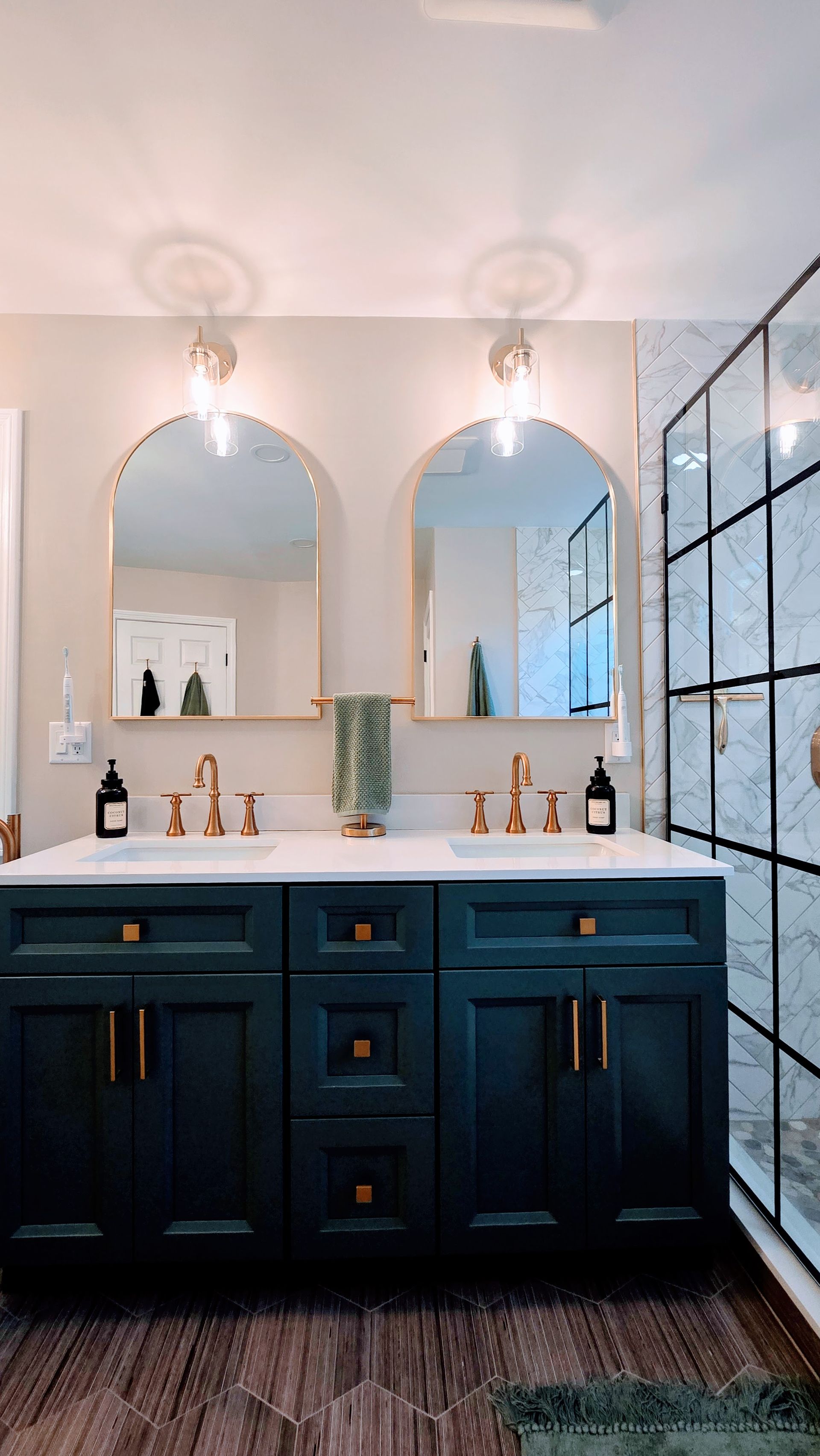 Dark blue bathroom vanity in Wolcott, CT  with gold fixtures, arched mirrors, and marble tile shower.