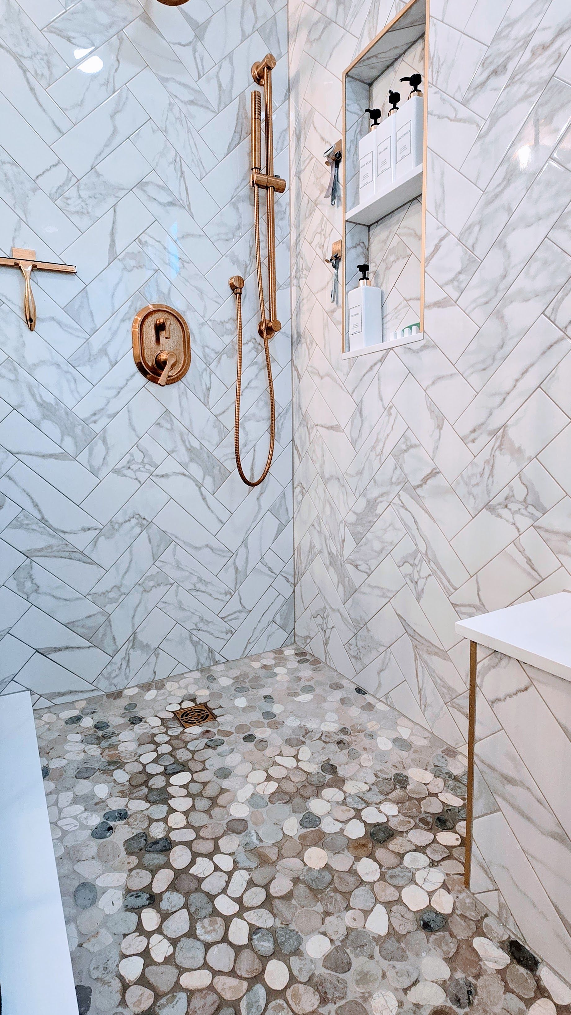Marble-tiled walk-in shower remodel in Wolcott, CT with gold fixtures, pebble floor, and recessed shelf with toiletries.