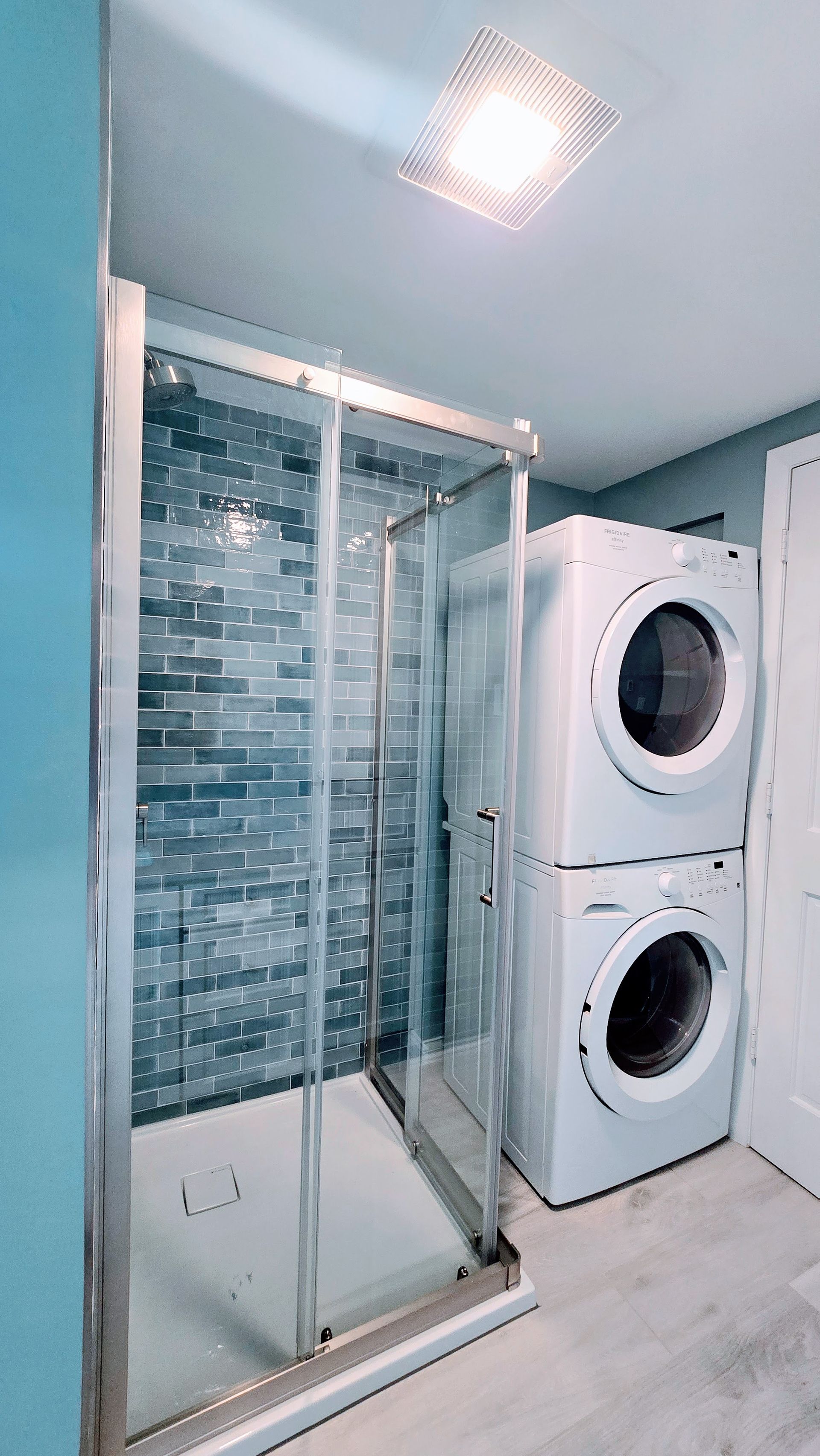 Bathroom remodel in Ashford, CT completed by First Choice Bathroom Remodeling with a glass shower, stacked washer/dryer, and blue-tiled wall. White floor and light fixture.