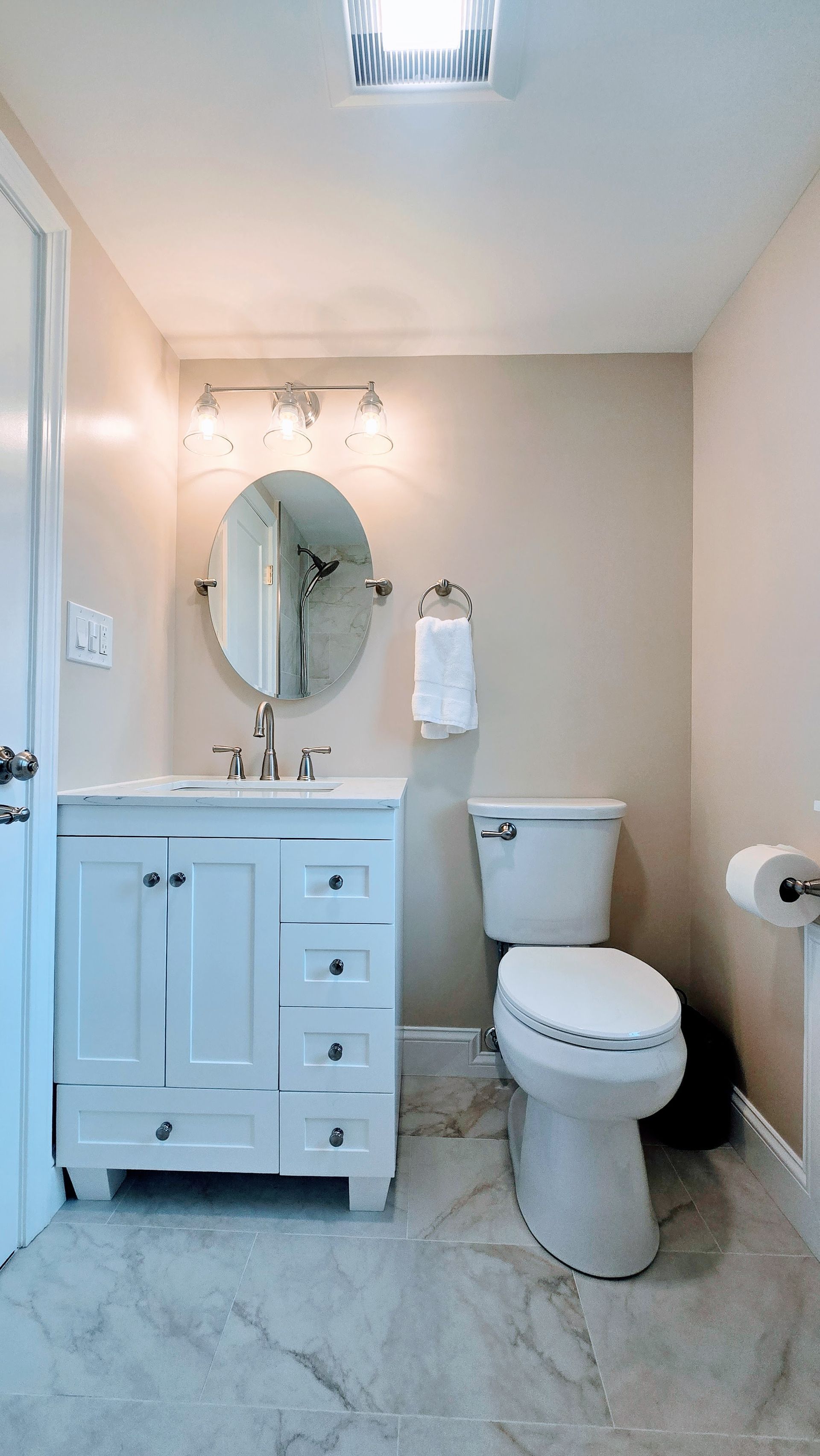 Small bathroom remodel in Southington, CT completed by First Choice Bathroom Remodeling with a with white vanity, toilet, and marble-look floor. Beige walls, oval mirror, and overhead light.