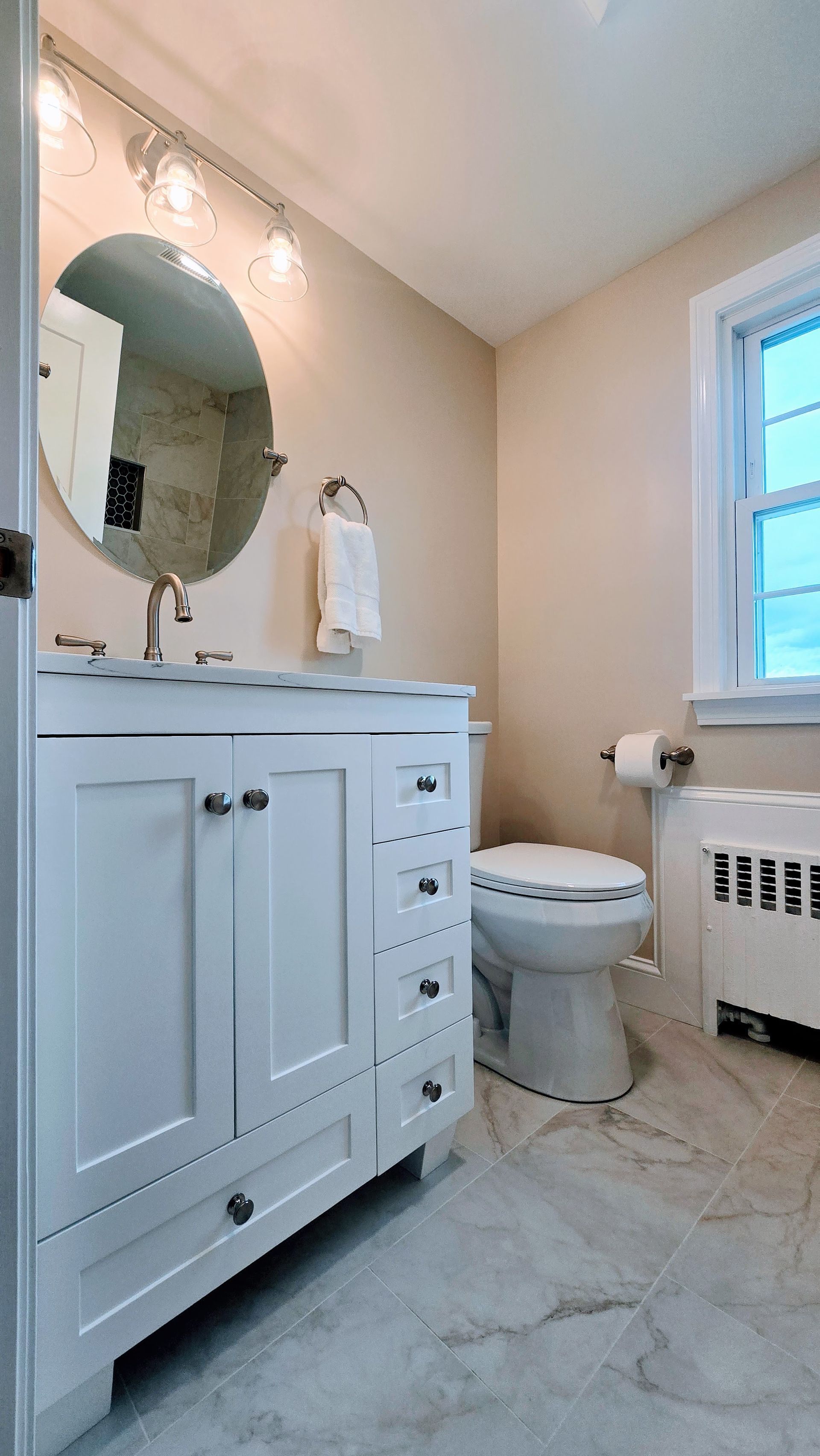 Small bathroom remodel in Southington, CT completed by First Choice Bathroom Remodeling with a  white vanity, round mirror, and toilet. Beige walls, window on the right.