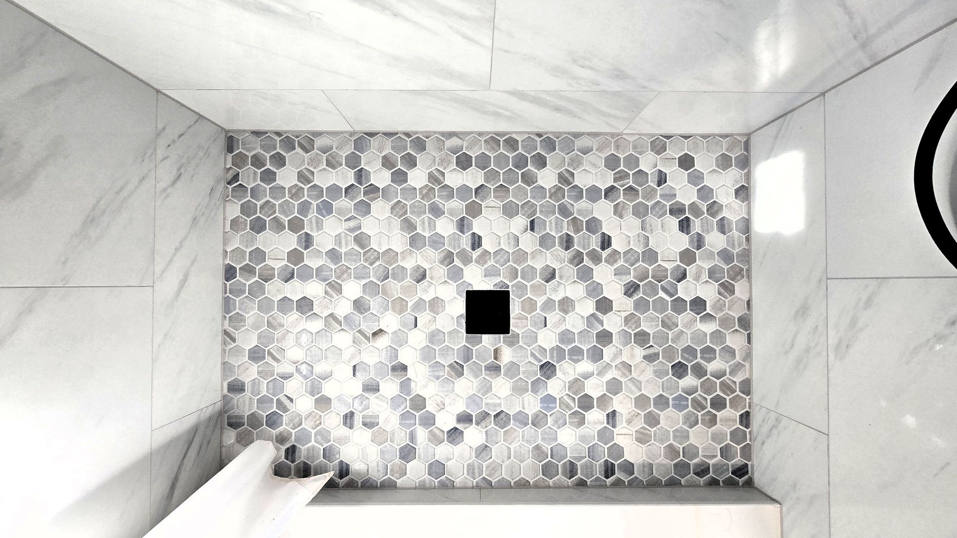 Shower remodel in Berlin, CT completed by First Choice Bathroom Remodeling with white marble walls and a hexagon tile accent wall with a black drain.