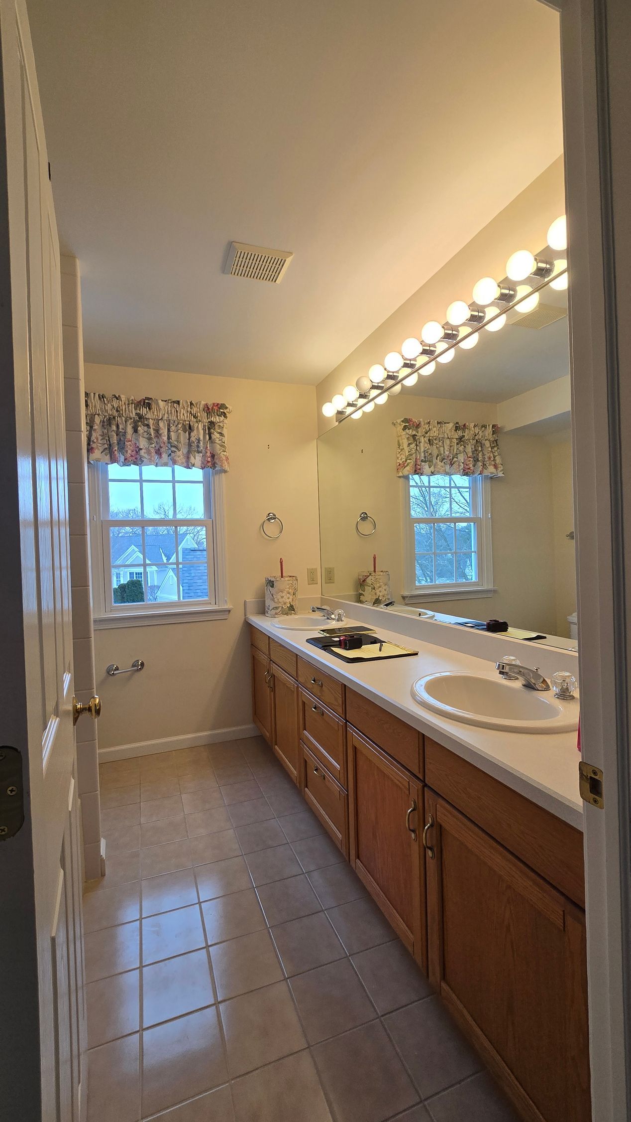 A brightly lit bathroom with wooden cabinets, a large wall mirror, white countertop, and a window with floral valances.