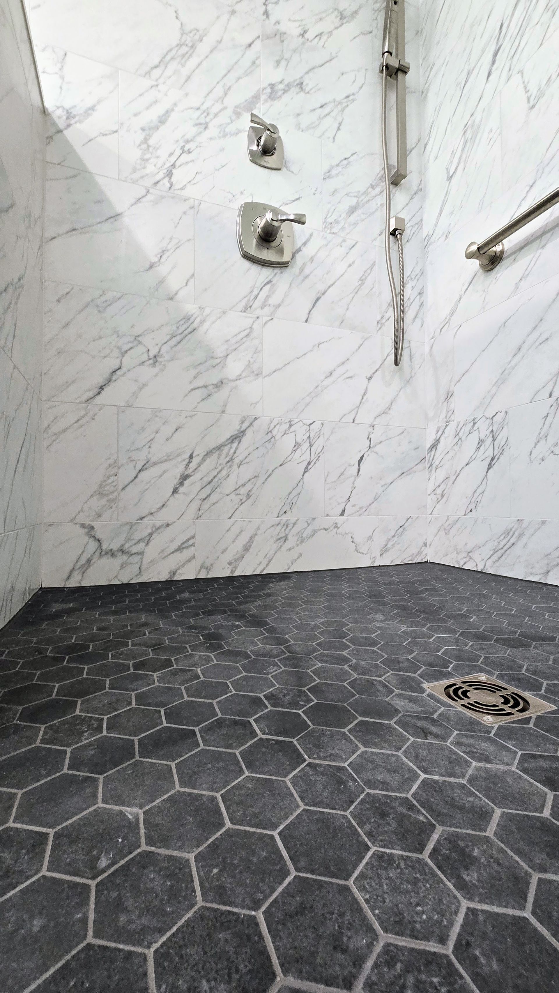 A shower stall with white marble-patterned walls in Glastonbury, CT, a charcoal hexagon tile floor, brushed-nickel fixtures, and a grab bar.