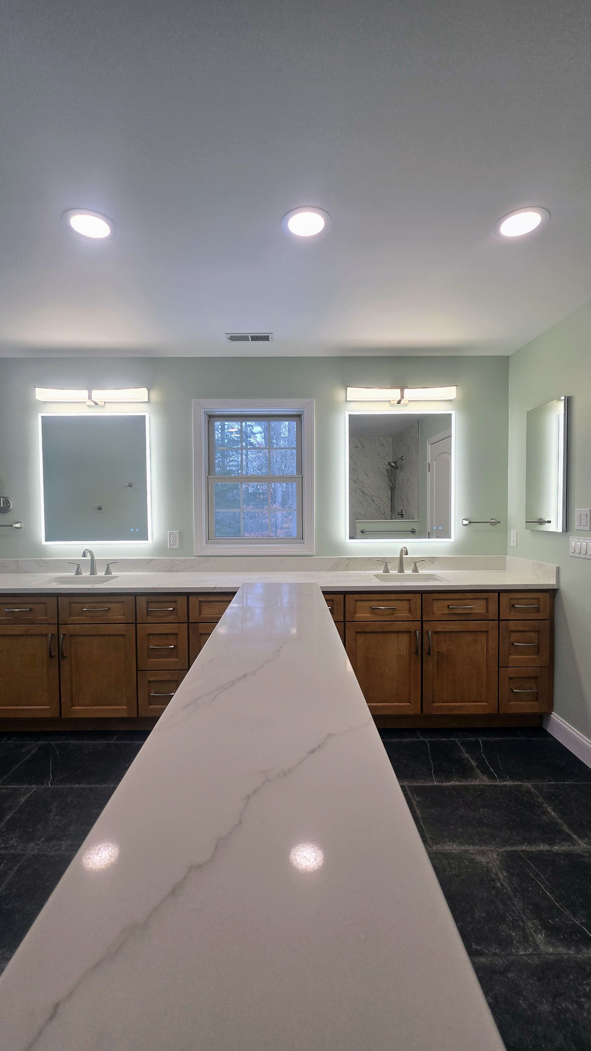 Bathroom vanity with two sinks, backlit mirrors in Glastonbury, CT, wooden cabinets, and a long marble-topped counter against a sage wall.