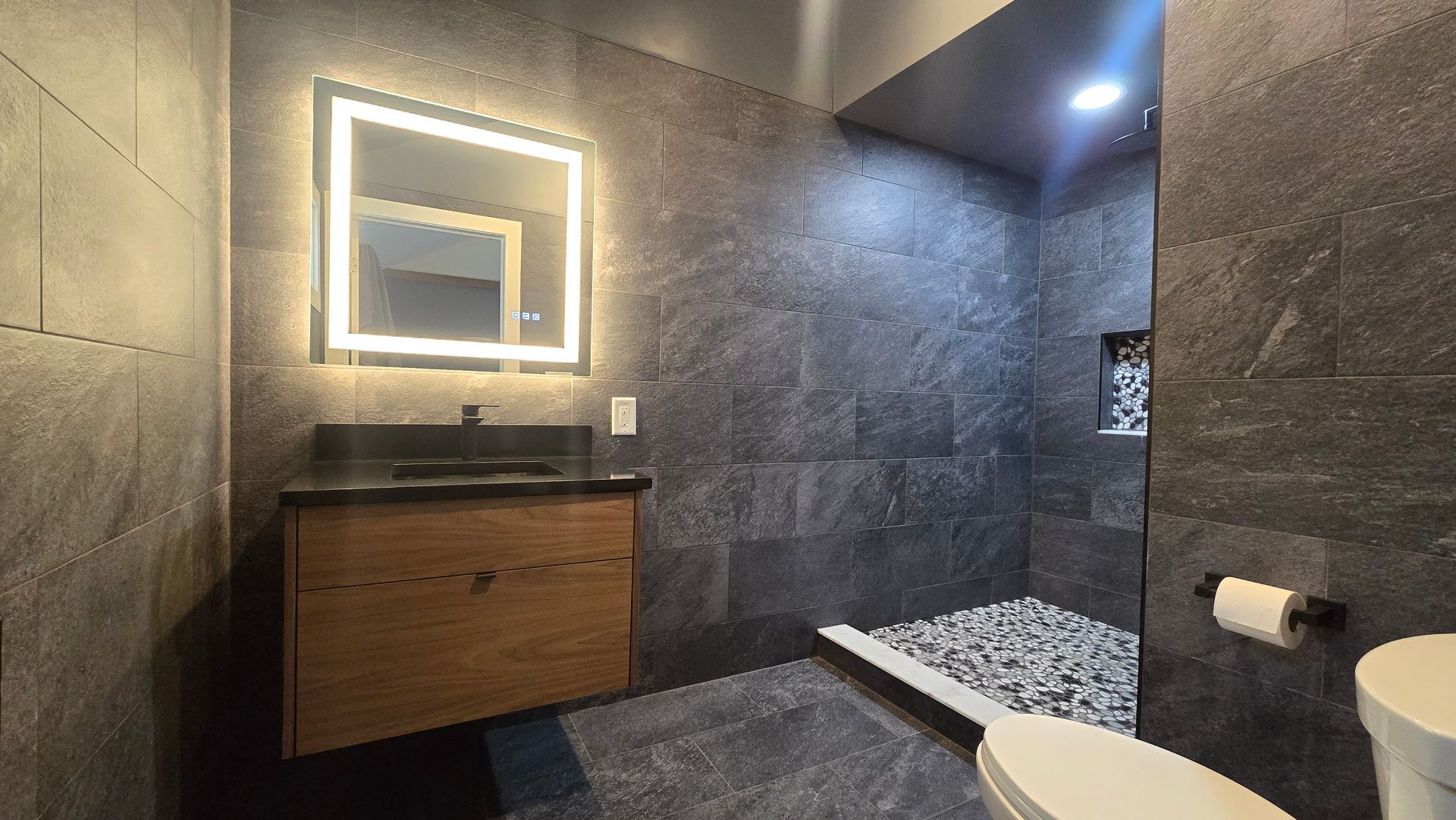 Modern bathroom with gray tile walls, illuminated mirror, wooden vanity, and shower.