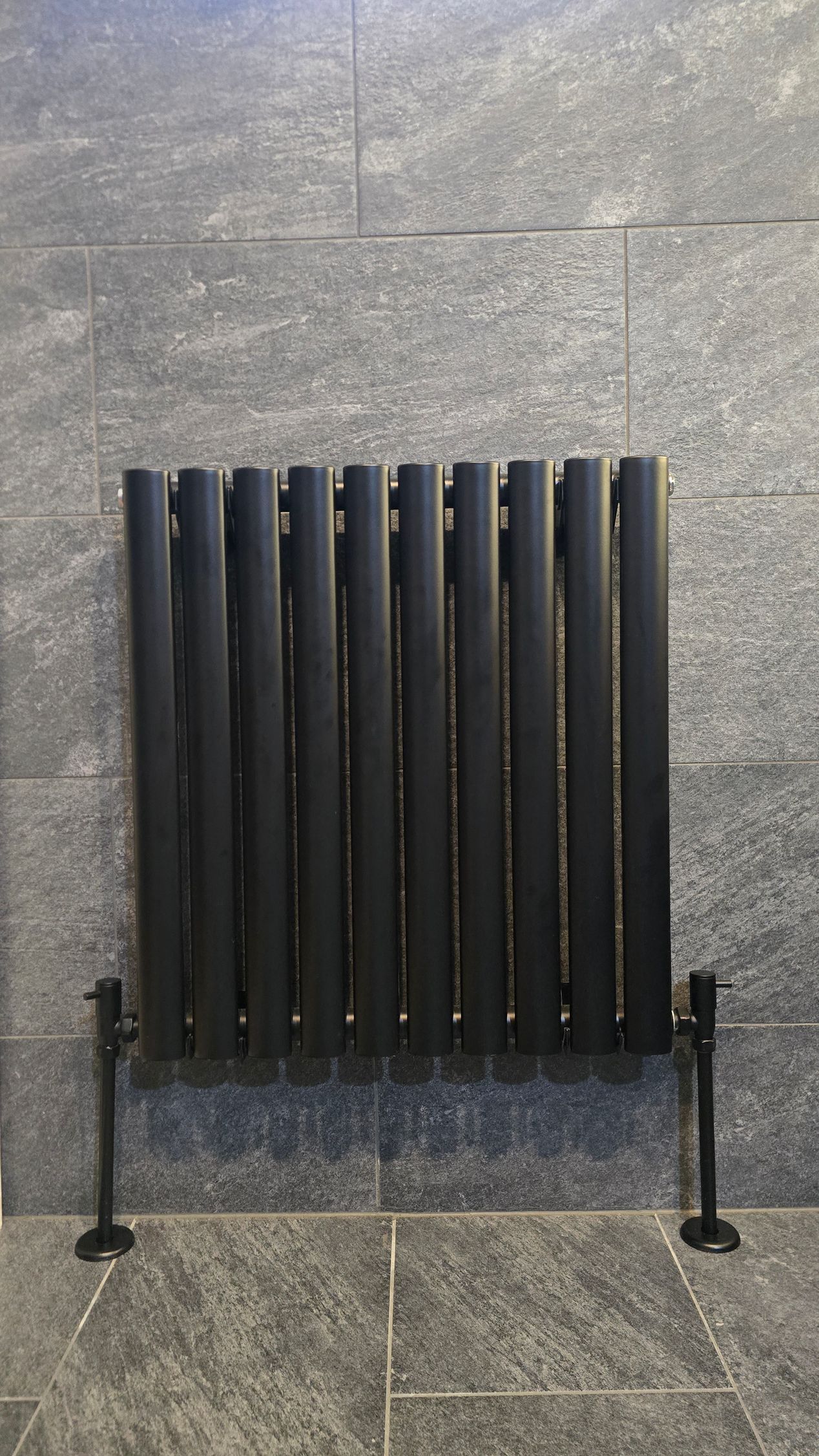 A black vertical-column radiator mounted on a dark, textured stone wall with two black floor-mounted support legs.
