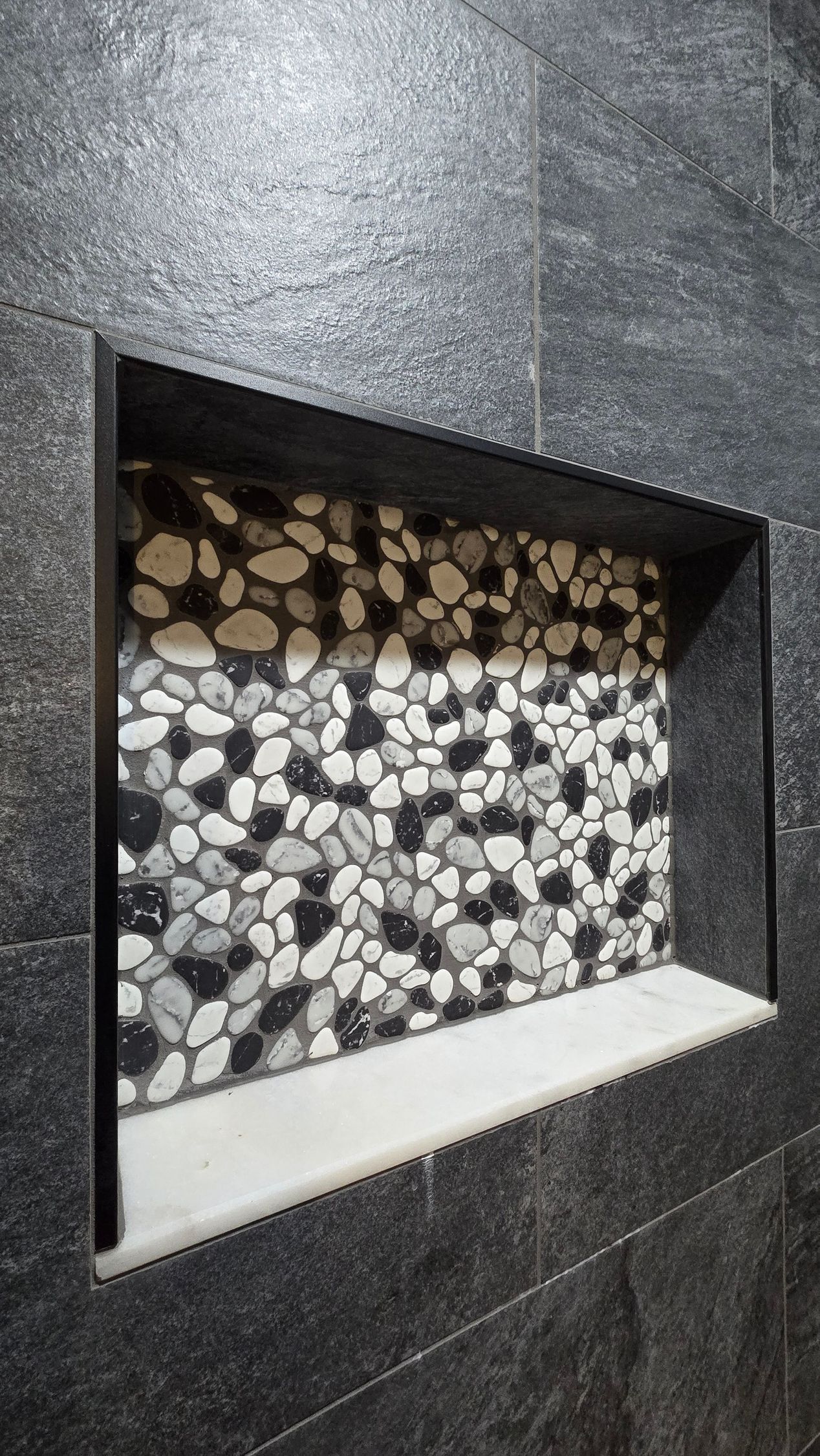 A rectangular shower niche in a dark grey tiled wall, featuring a back wall of white, grey, and black river stones.