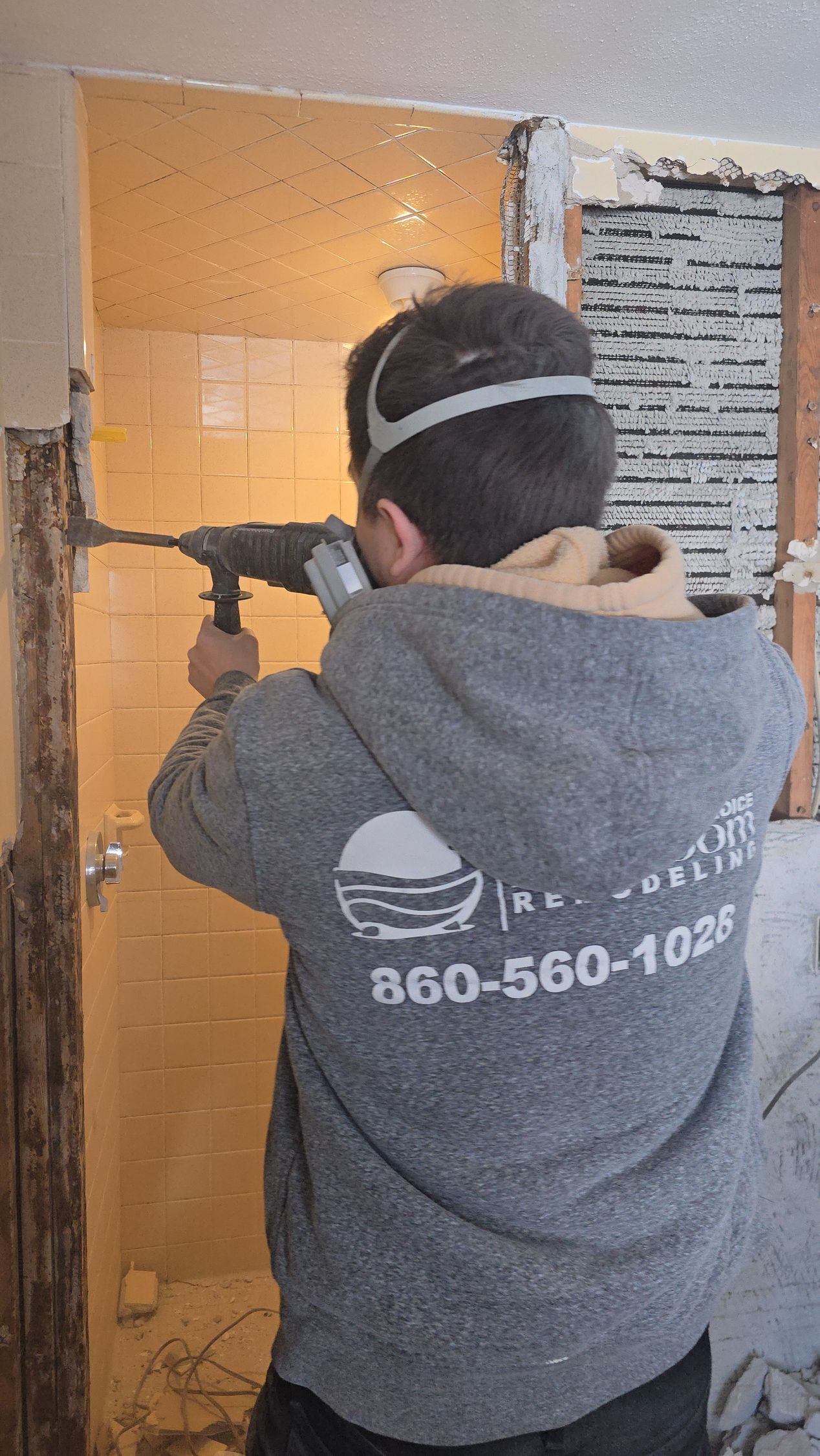 First Choice Bathroom Remodeling team member in Simsbury, CT in a gray hoodie wearing a protective mask uses a power tool to demolish a wall in a room under renovation.