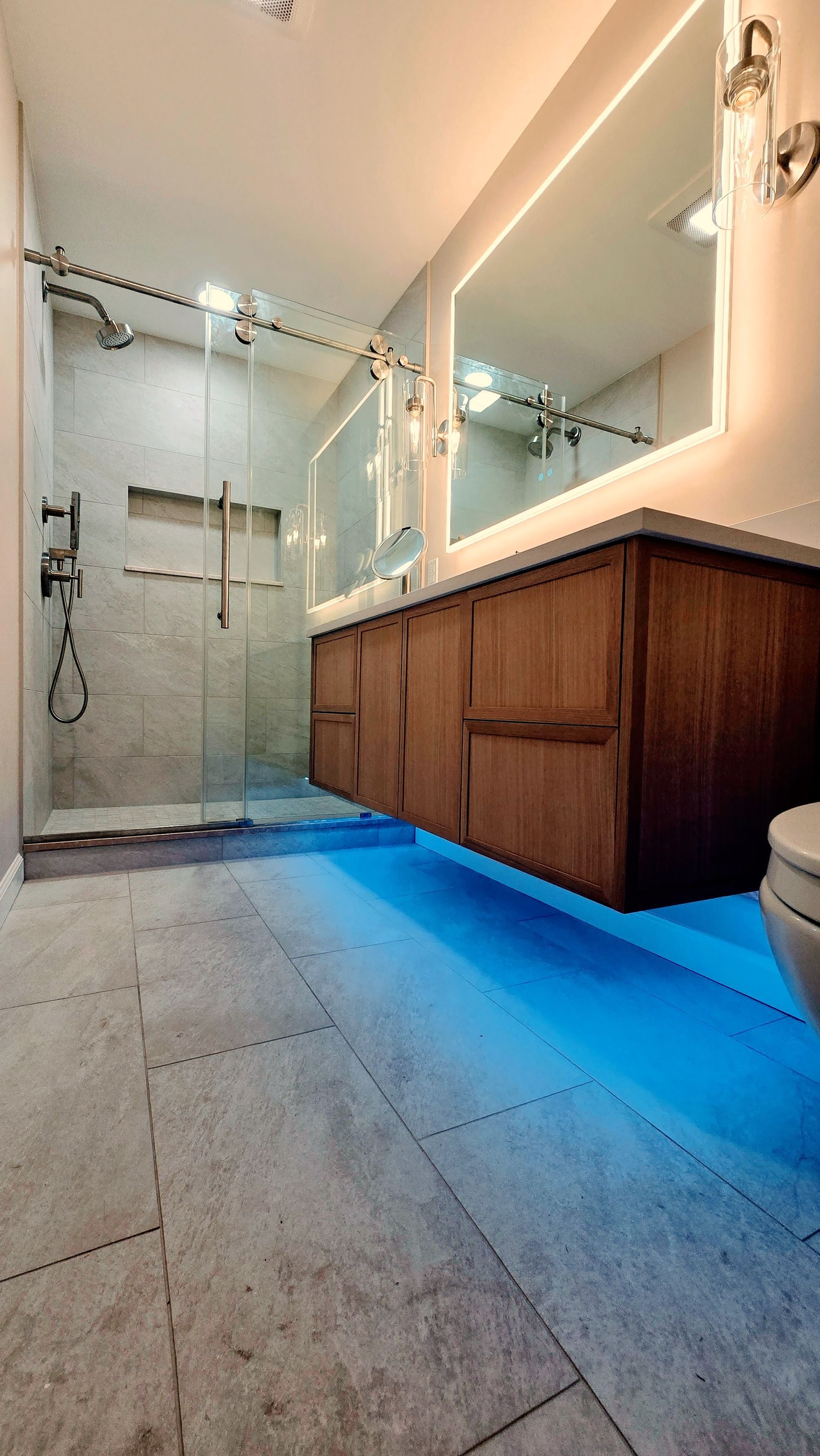 A modern bathroom remodel in Simsbury, CT featuring a floating wooden vanity with under-cabinet blue lighting, stone tiling, and a glass shower.