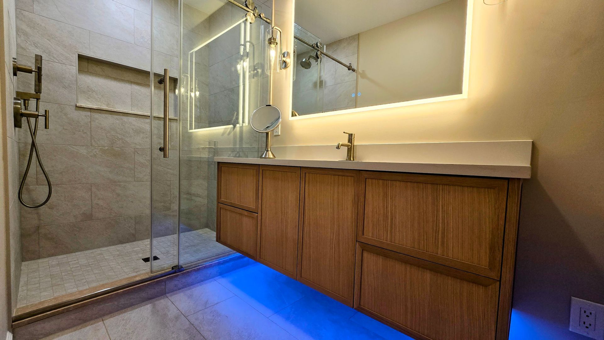Modern bathroom featuring a wood floating vanity, illuminated mirror, walk-in glass shower, and blue ambient floor lighting.