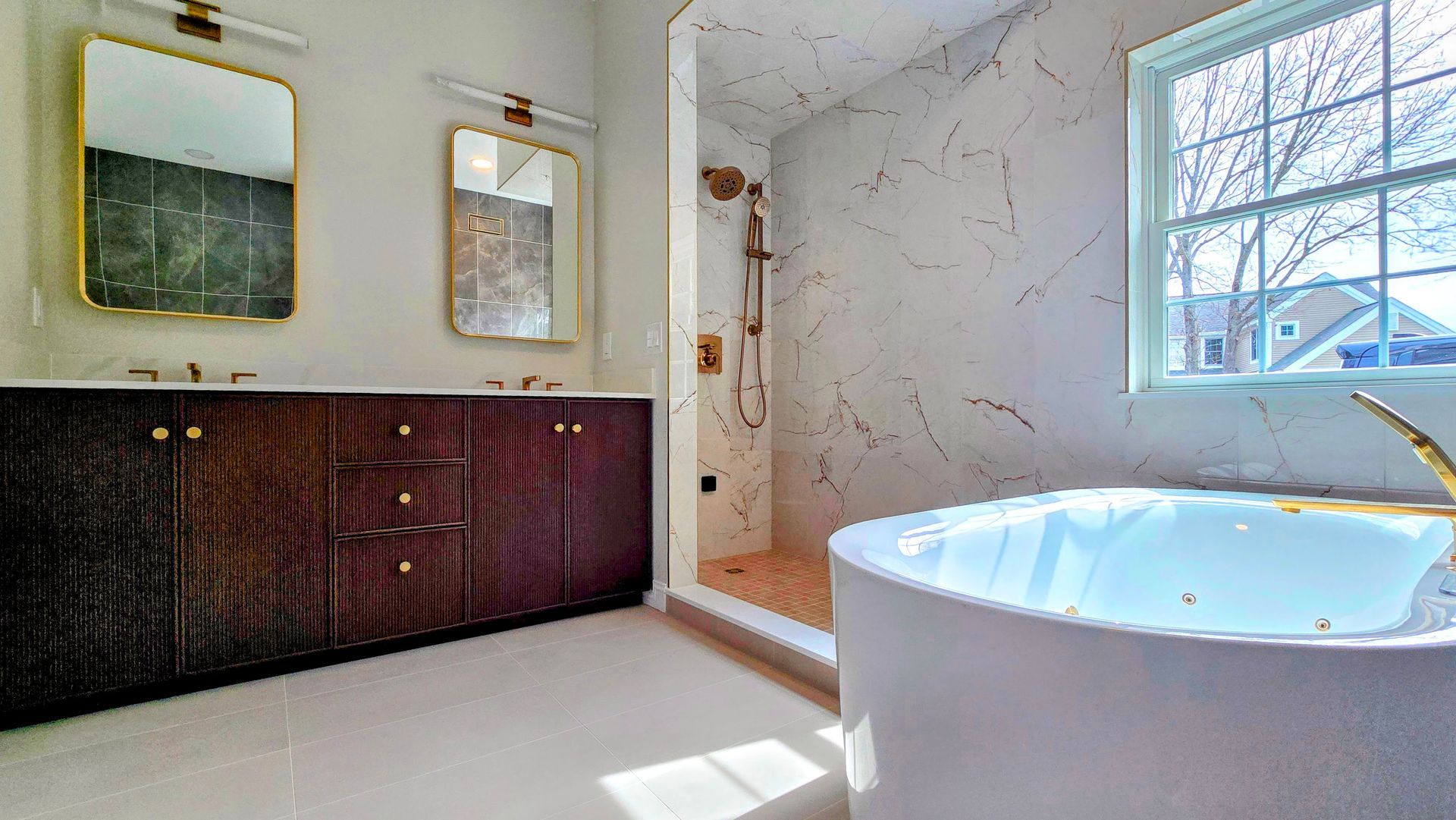 A modern bathroom in Cromwell, CT with a dark wood vanity, gold mirrors, a white soaking tub, and a walk-in shower with marble-look tiles.