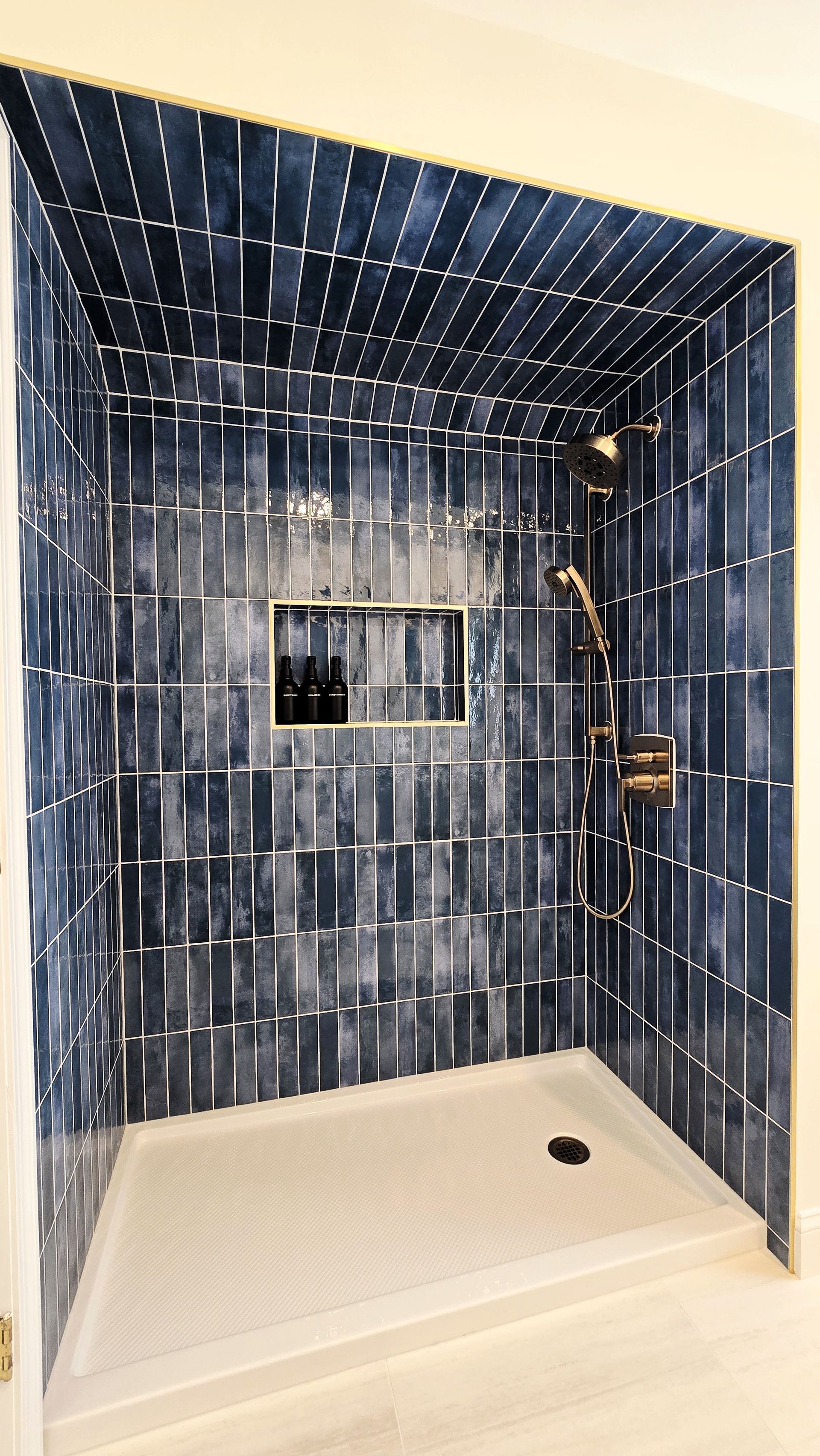 A modern walk-in shower in Cromwell, CT with dark blue vertical tiles, a built-in recessed shelf, and gold hardware.