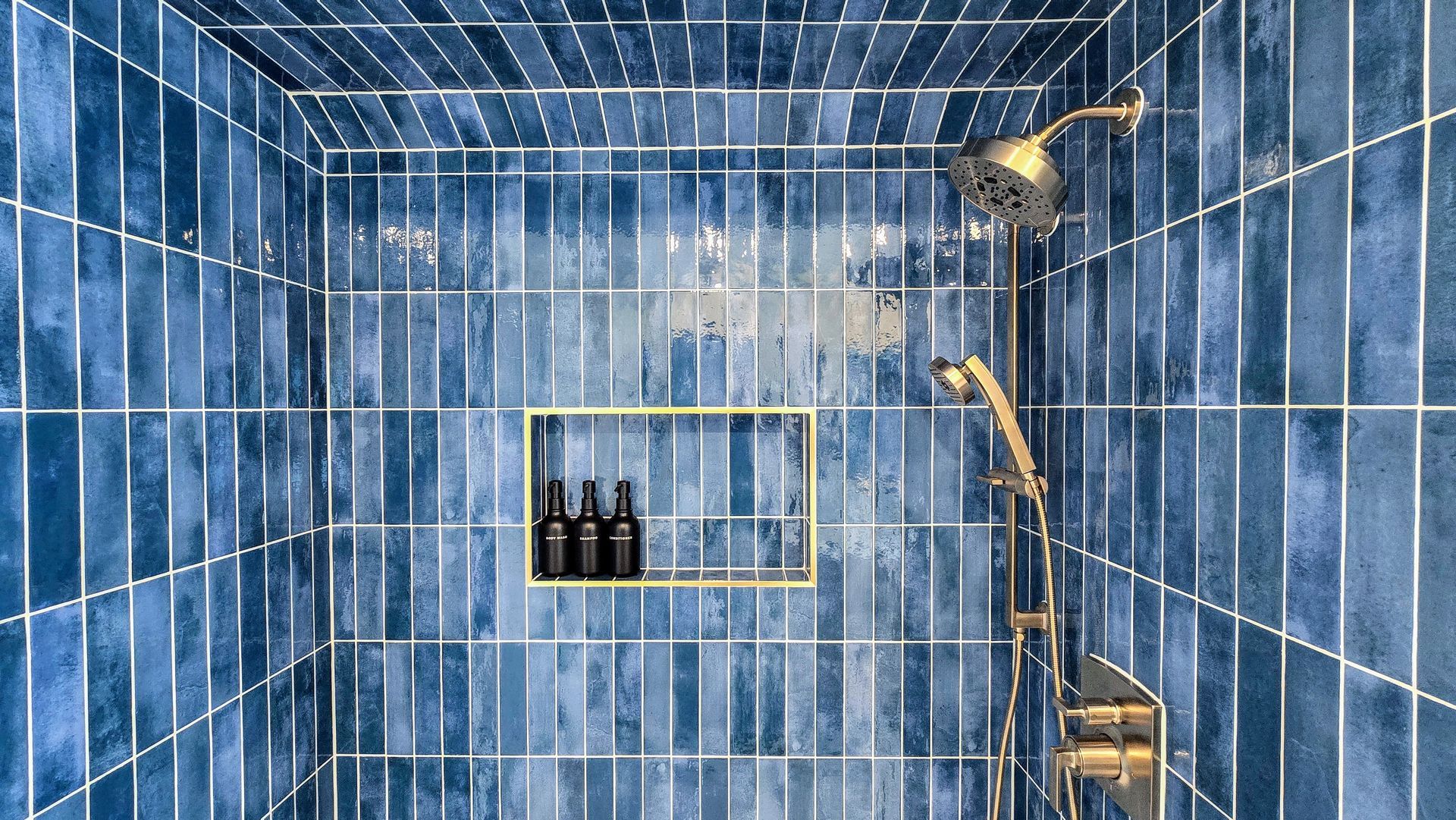 A walk-in shower in Cromwell, CT with vertical, mottled blue tiles, gold fixtures, and a recessed wall niche holding three dark bottles.