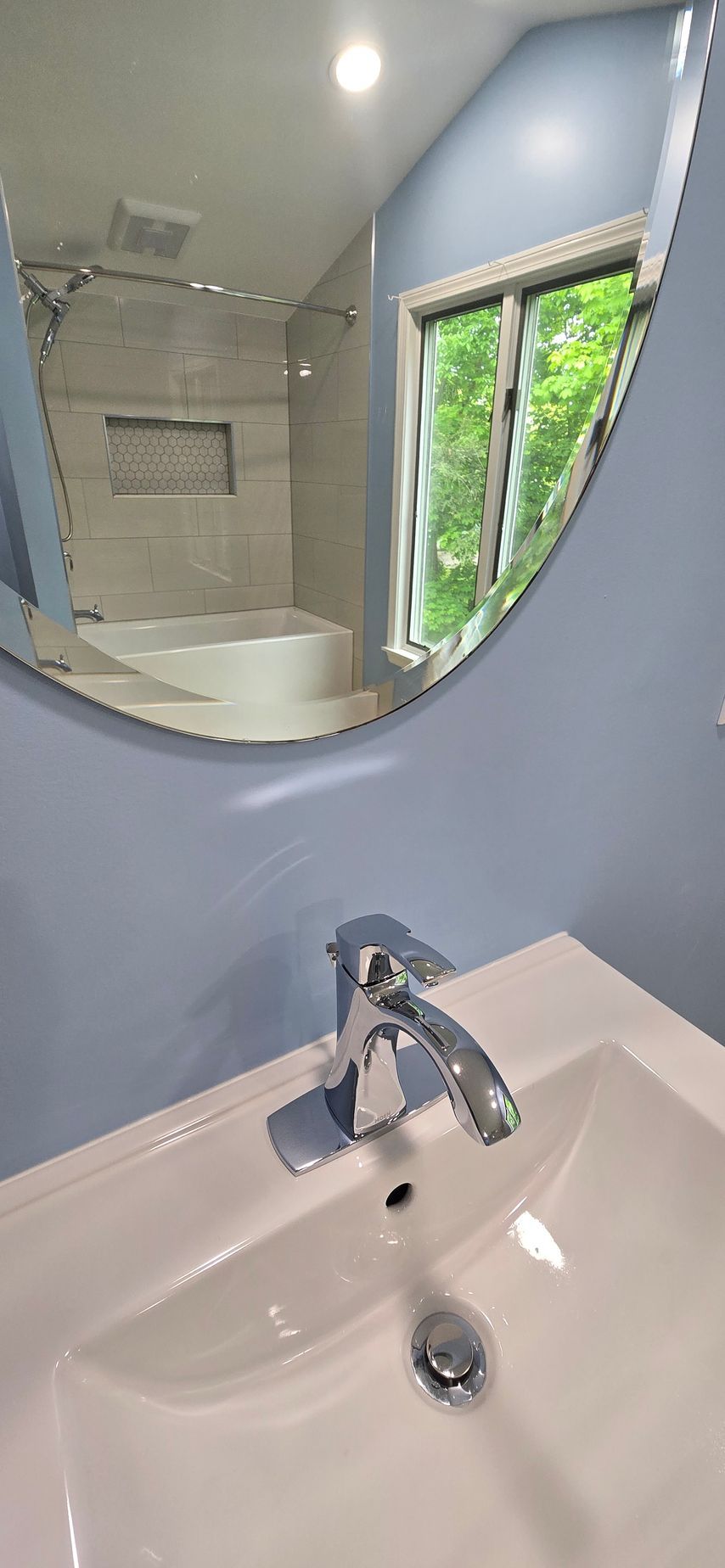 Bathroom sink and mirror reflecting a bathtub, tiled shower, and window with greenery outside.
