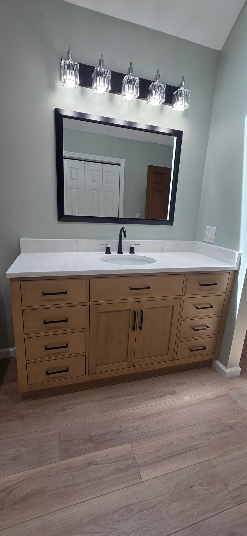 A bathroom vanity with a light wood cabinet featuring black hardware, a white countertop with a sink and faucet, a large black-framed mirror, and a black light fixture with glass shades above it. The walls are painted a soft green, and there is light wood flooring.
