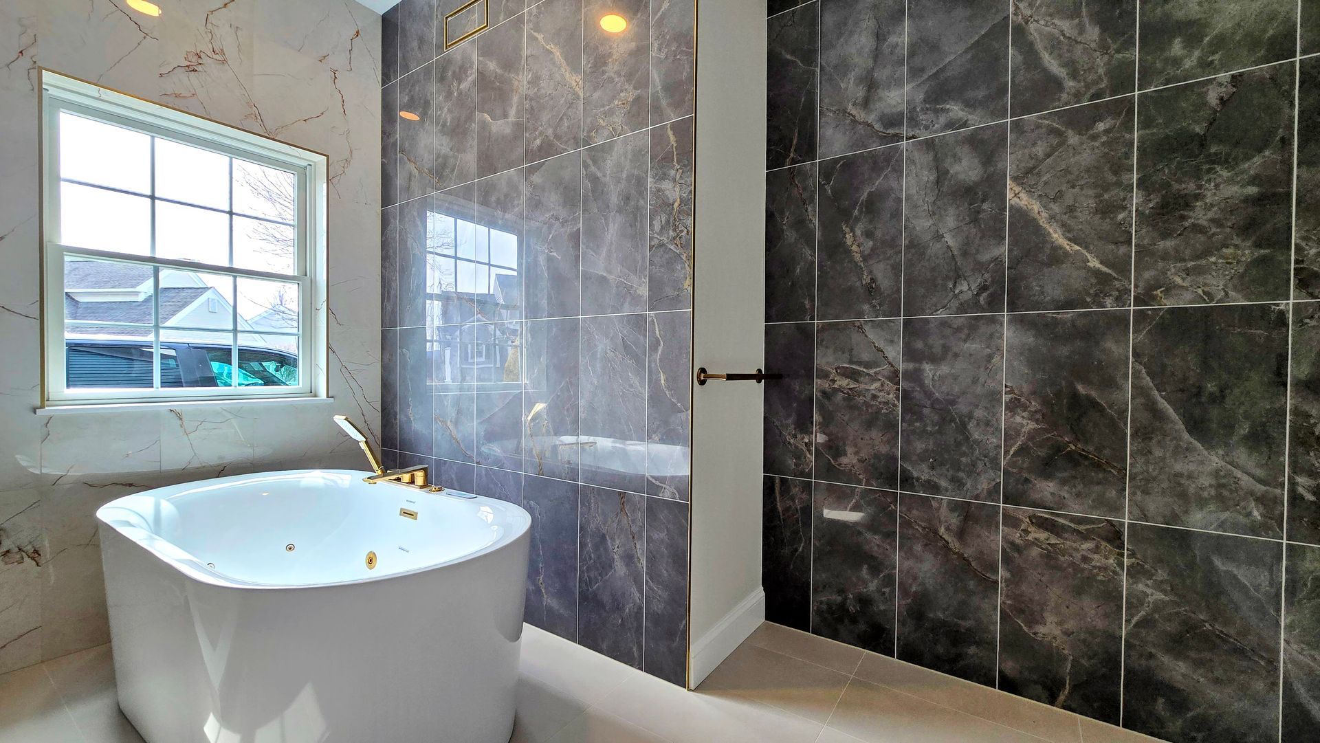 A white soaking tub in Cromwell, CT sits by a window next to a large shower wall covered in dark, metallic-accented marble tiles.
